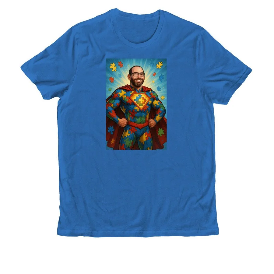 Blue T-shirt featuring a graphic of a man smiling, dressed as a superhero with a puzzle-piece costume, superhero emblem on chest, and a cape, surrounded by puzzle pieces.