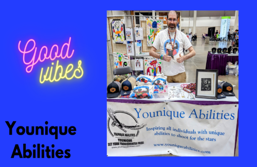 A man standing behind a table at a Younique Abilities booth, displaying hats and framed photos on a banner that reads 'Younique Abilities,' during an event with colorful decorations and other booths in the background.