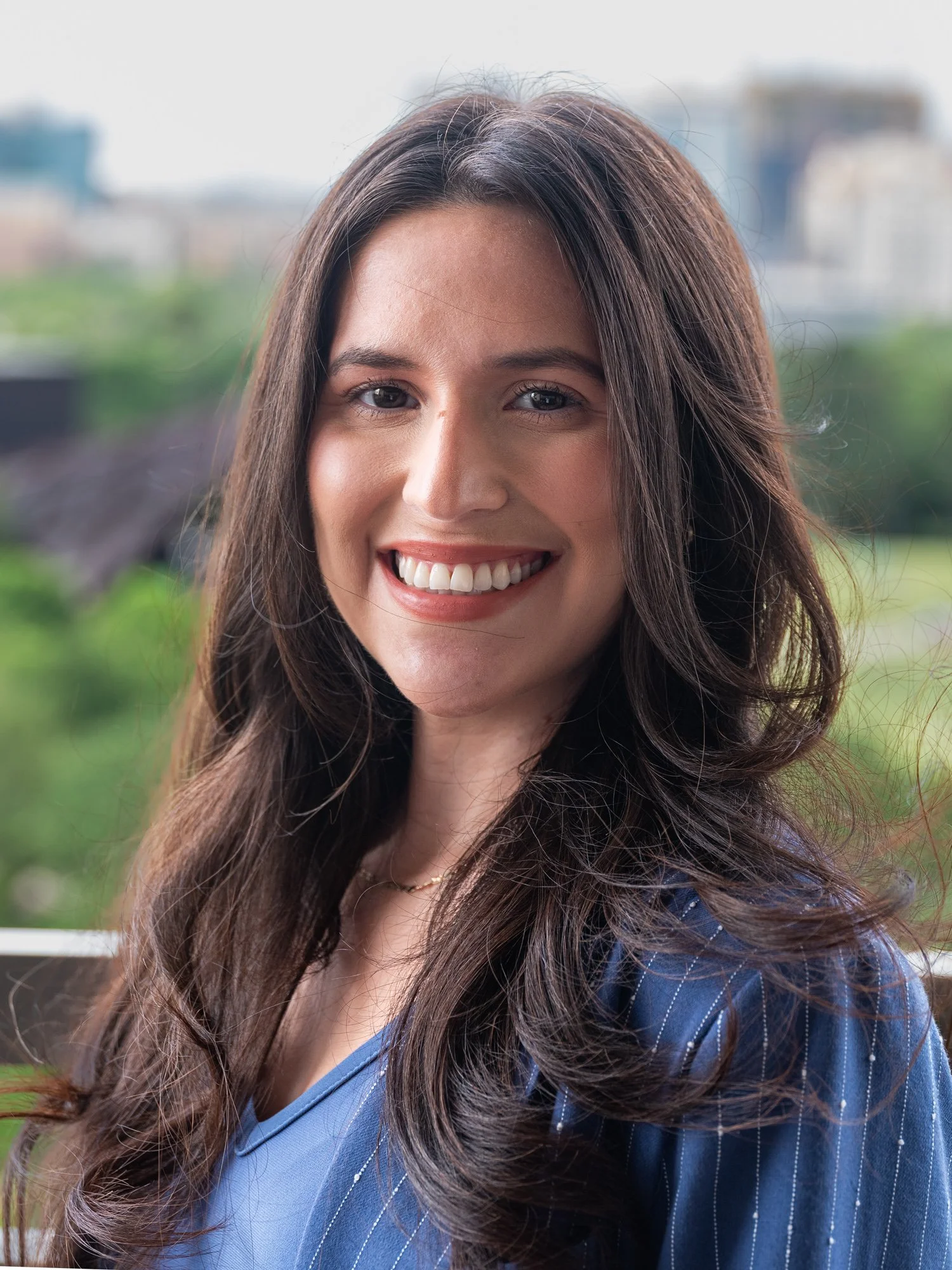 Close-up of a smiling woman with long brown hair, wearing a blue shirt, outdoors with blurred buildings and greenery in the background.