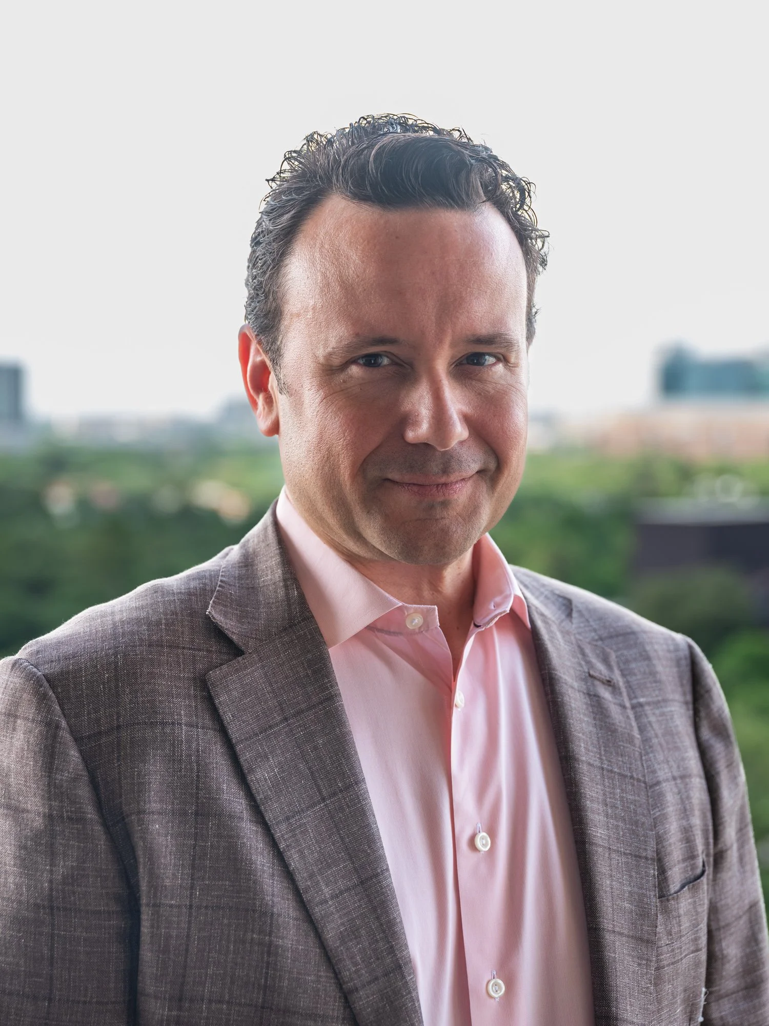 A man with dark, wavy hair styled back, wearing a light pink dress shirt and a brown checkered blazer, standing outdoors with a blurred cityscape and greenery in the background.