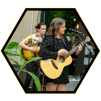 Women performing live music outdoors, one playing guitar and singing into a microphone, another playing guitar in the background, with green plants around.