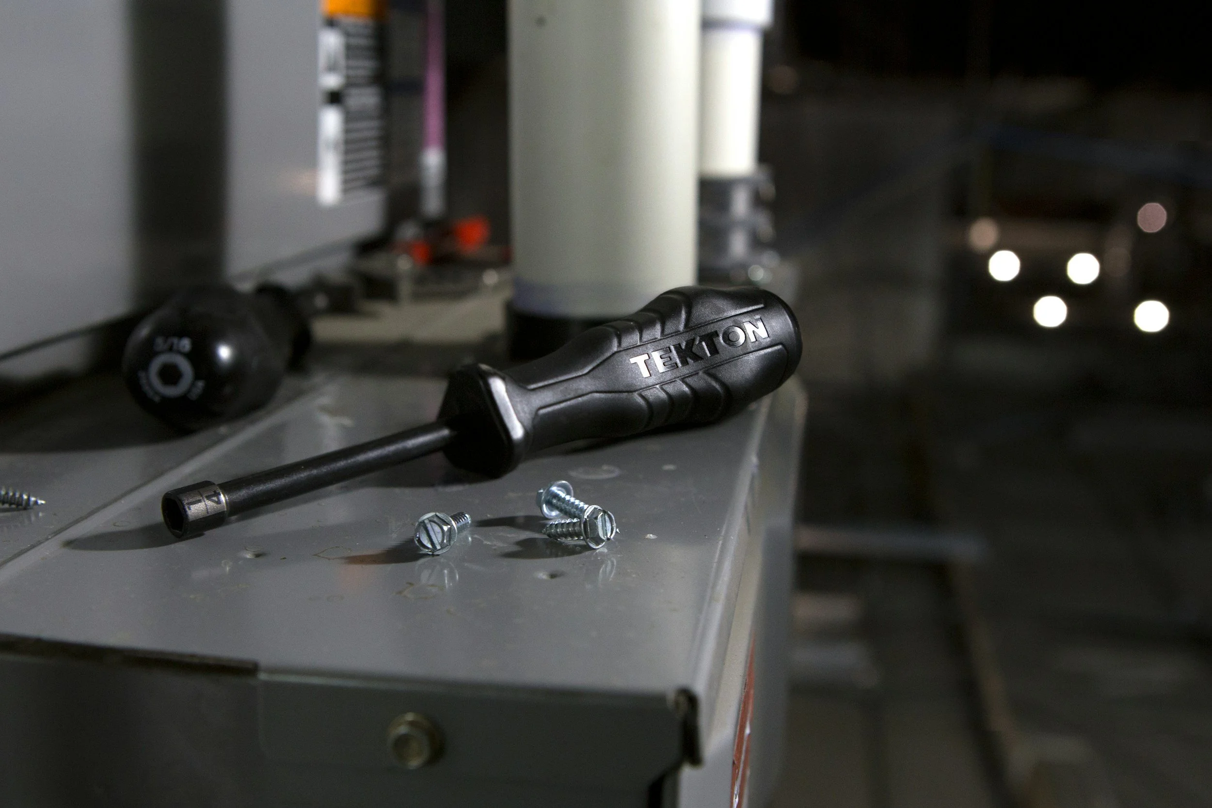 A screwdriver and several screws on a metal workbench in a garage or workshop, with a car's headlights visible in the background at night.