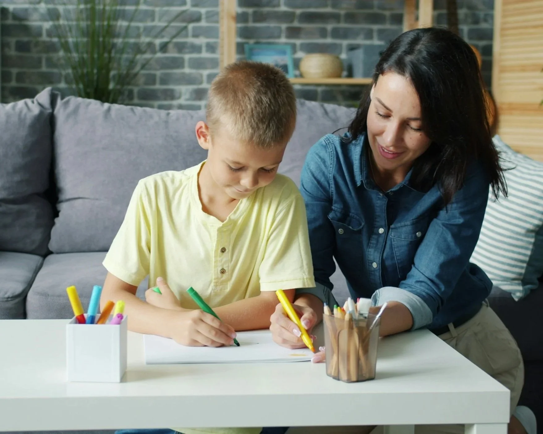 A woman and a young boy drawing together at a white table with colored markers in a cozy indoor setting.