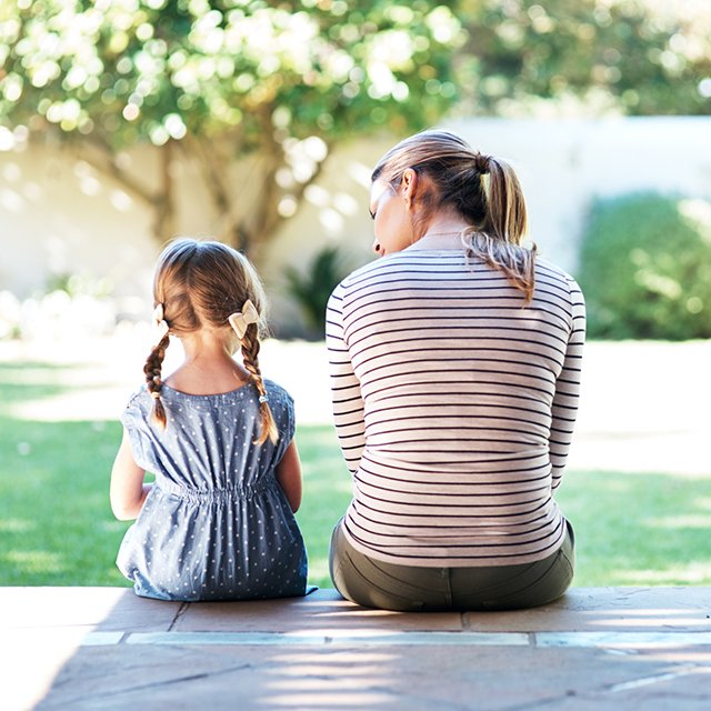 A woman and a young girl sitting on a park bench, facing away, with trees in the background on a sunny day.