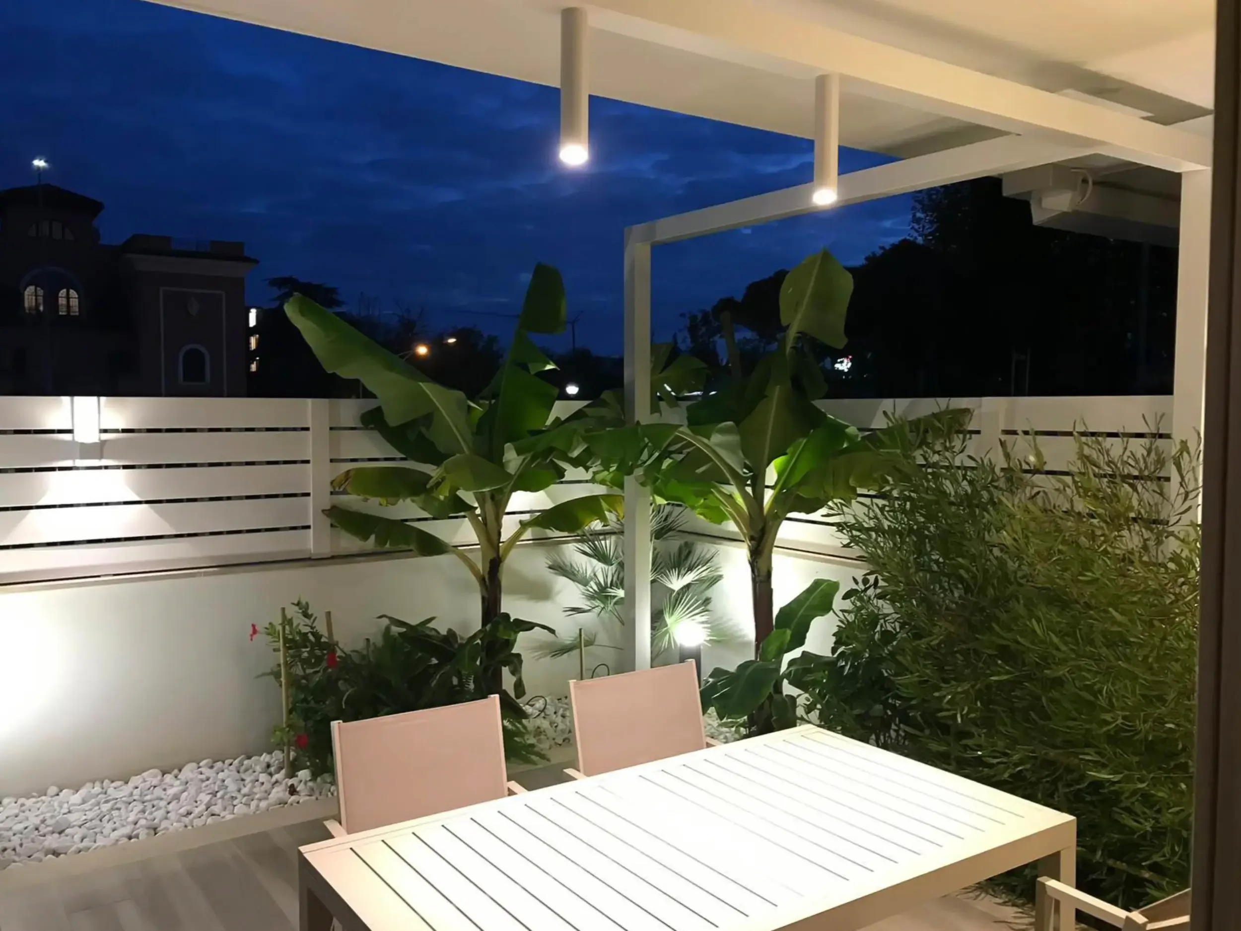 Galleria: Un tavolo esterno circondato da pannelli a griglia con luci accese dopo il tramonto.