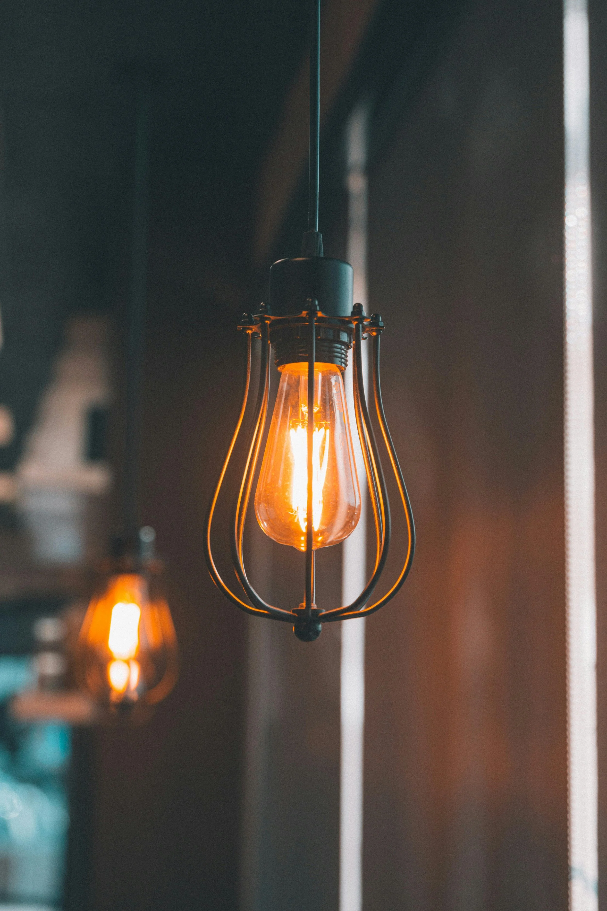 An Edison-style filament light bulb hanging from a ceiling, turned on and emitting a warm glow, with a second blurred light bulb in the background.