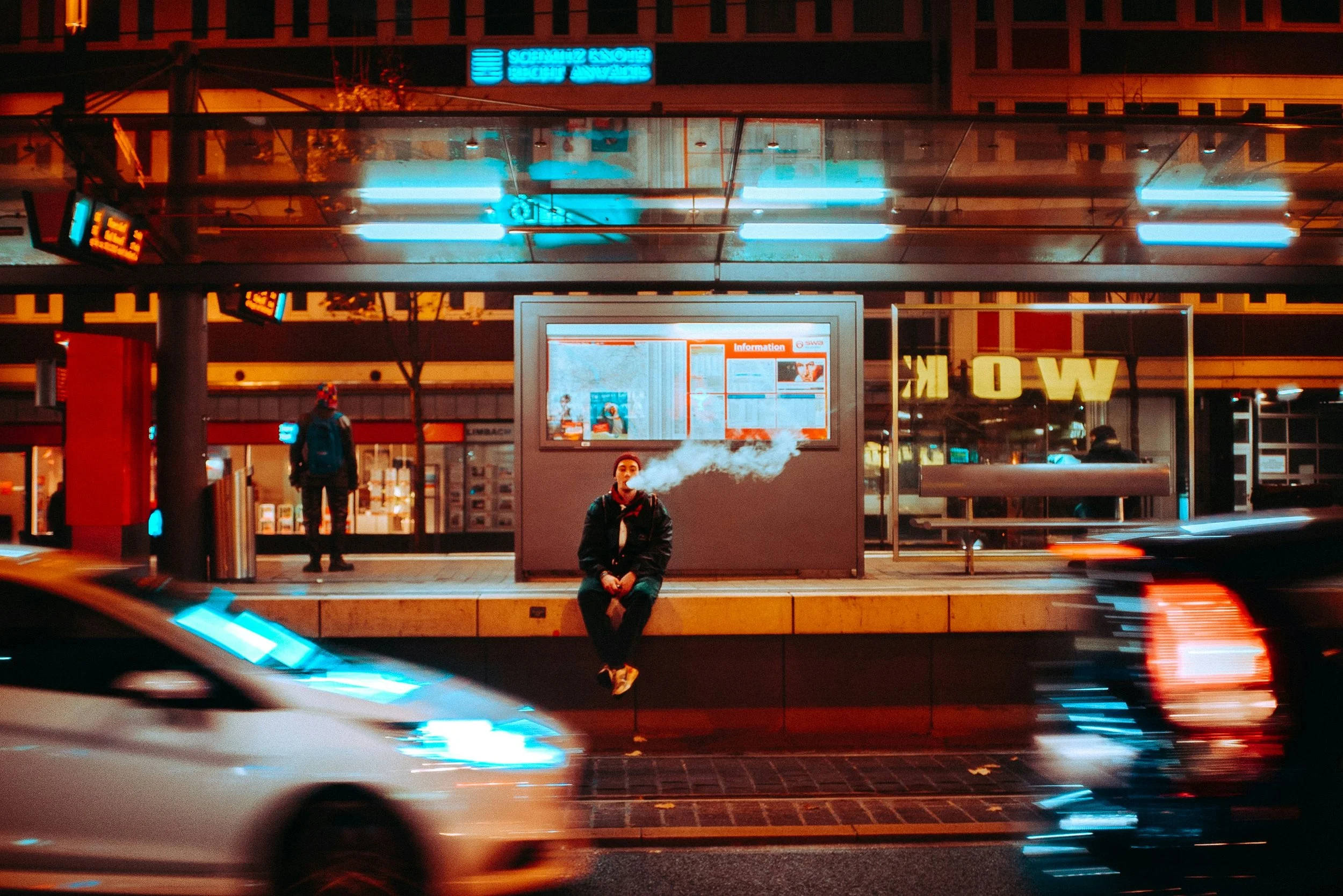 A person sitting on a bench in a city street at night, blowing smoke, with moving cars in the foreground and illuminated signs in the background.