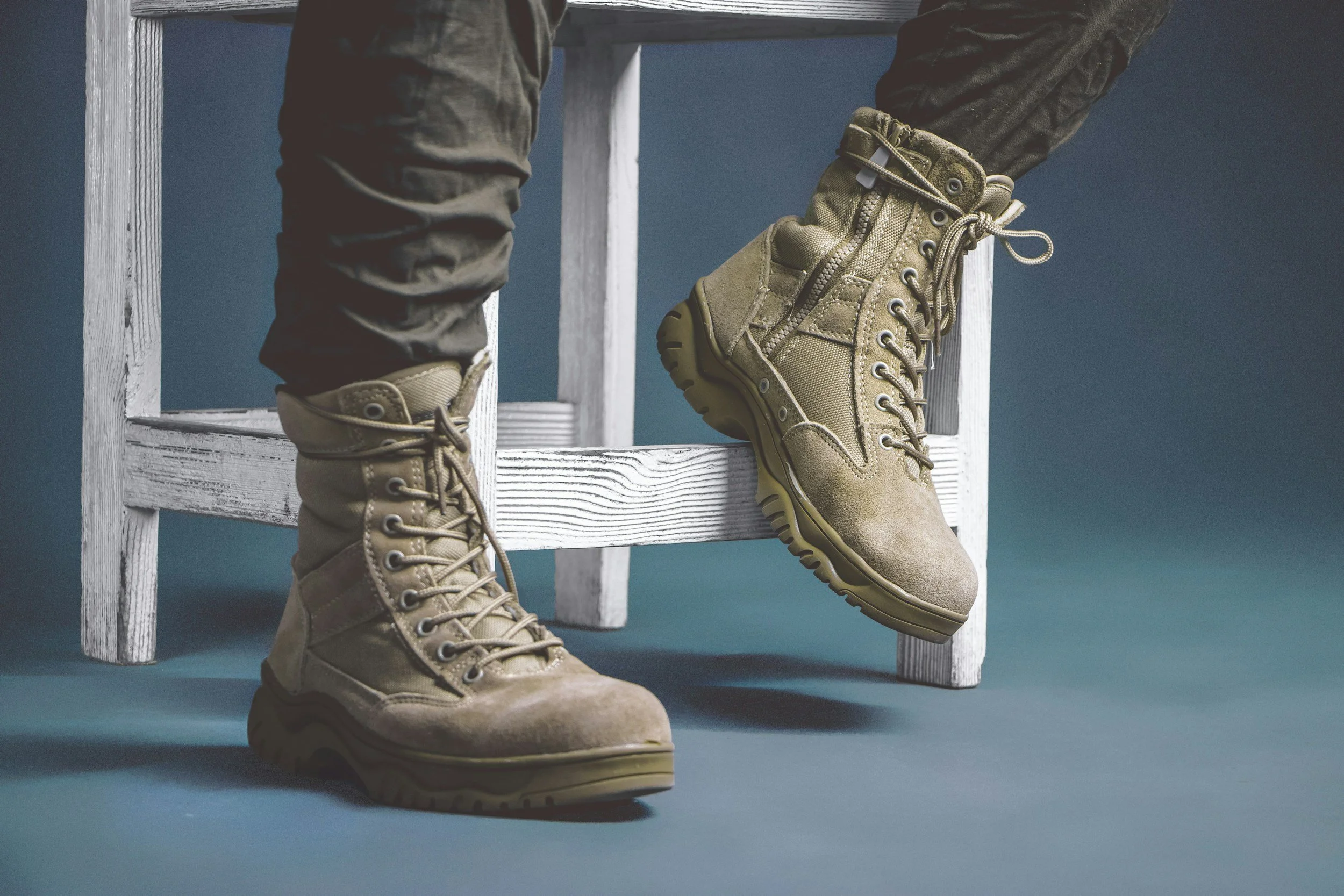 Close-up of tan tactical boots worn by a person sitting on a white wooden chair, with a dark teal background.