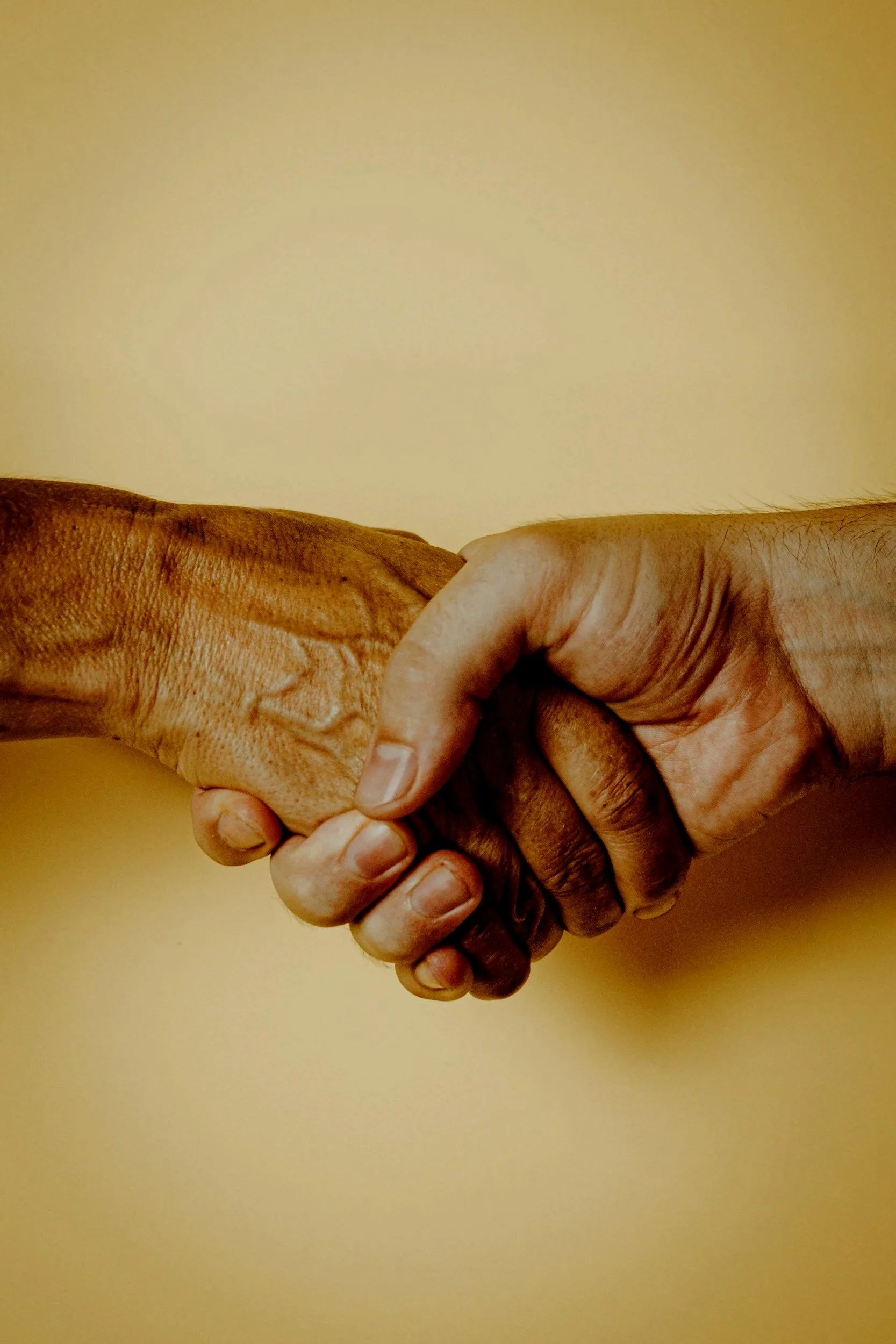 Two hands clasped in a handshake, with one hand darker-skinned and the other lighter-skinned, against a plain yellow background.