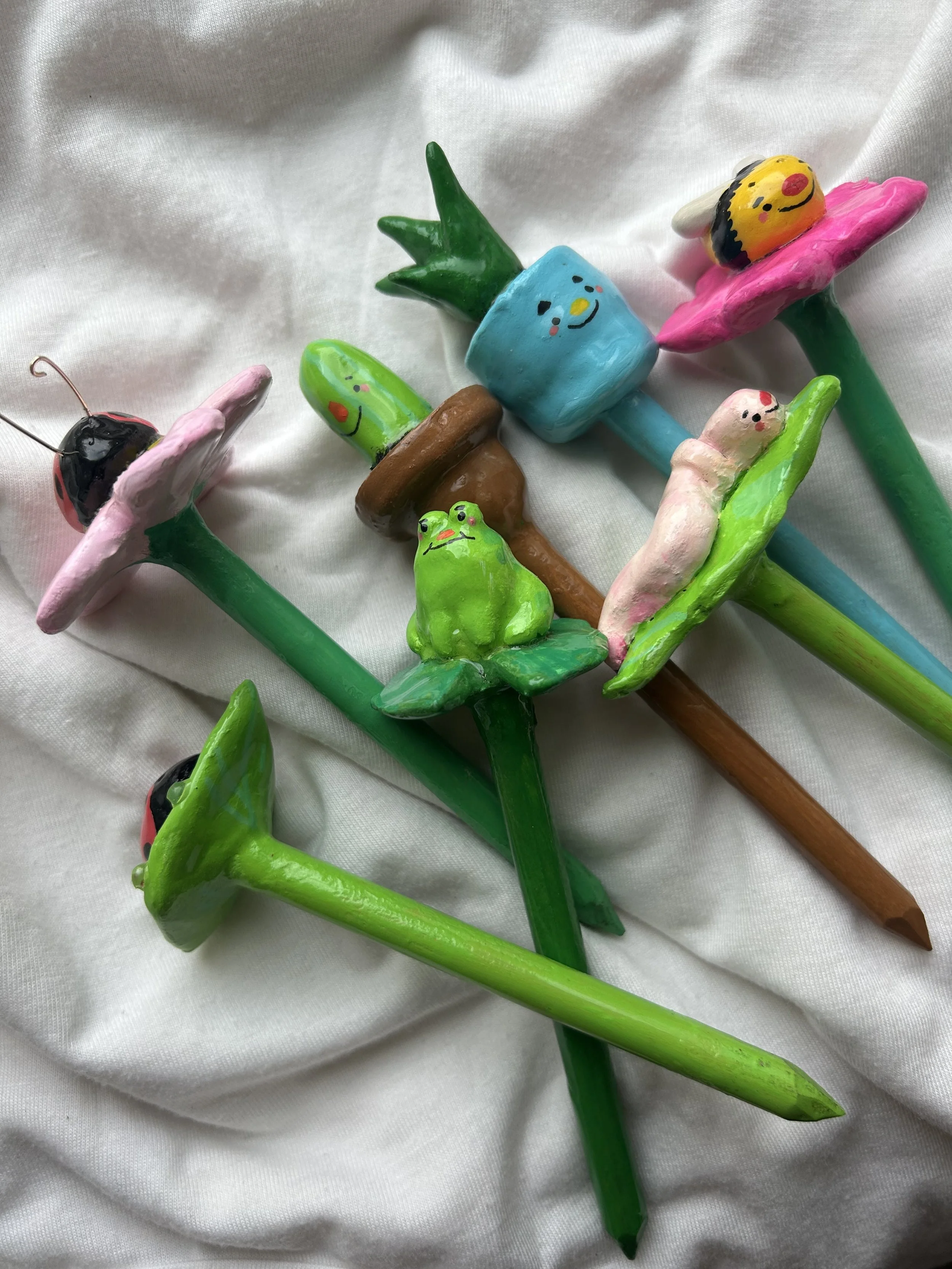 Colorful handmade clay garden stakes with whimsical characters, including ladybugs, frogs, and smiling plants, placed on a white fabric background.