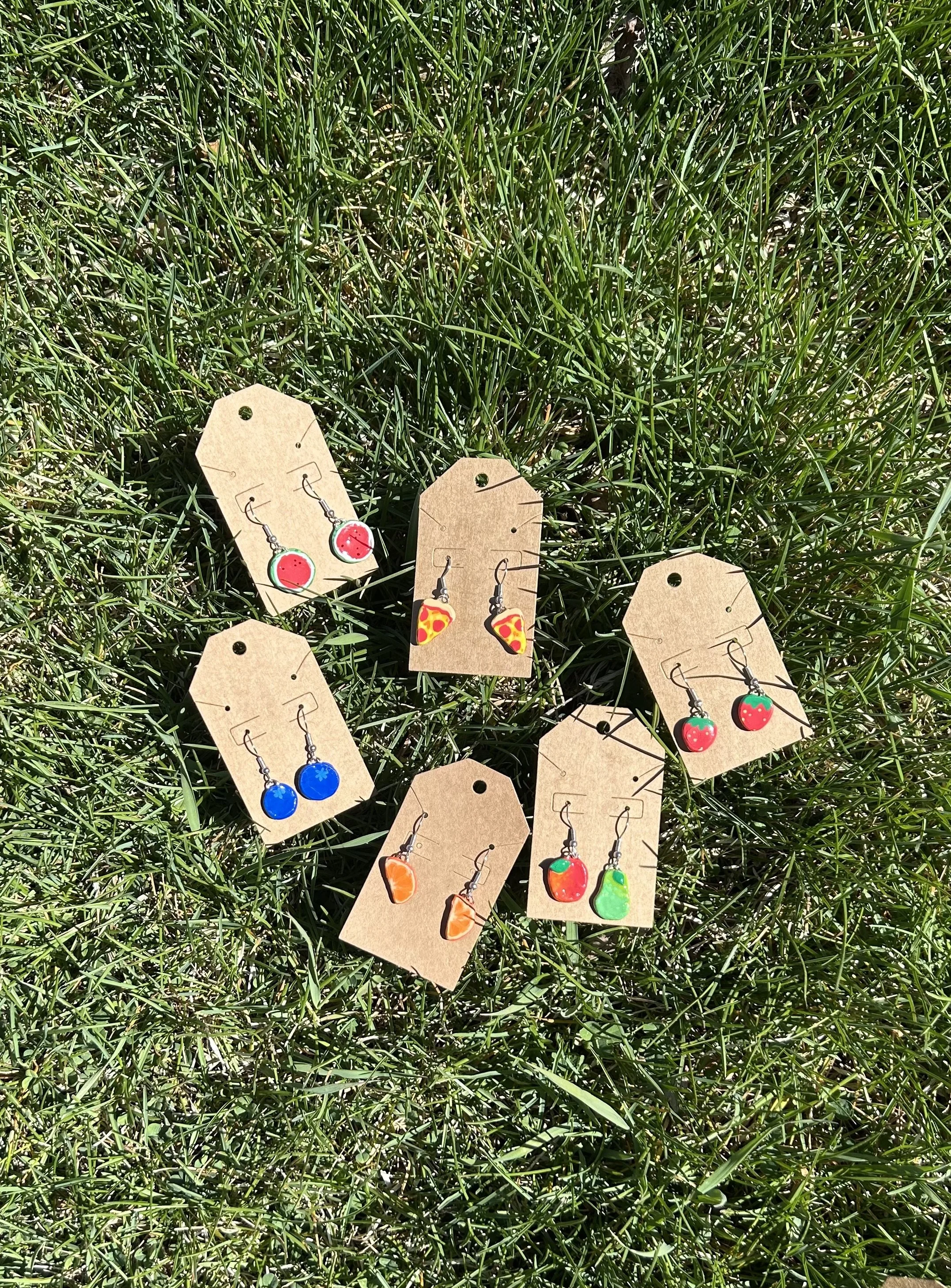 Colorful fruit-shaped earrings on tan paper tags laid on green grass.