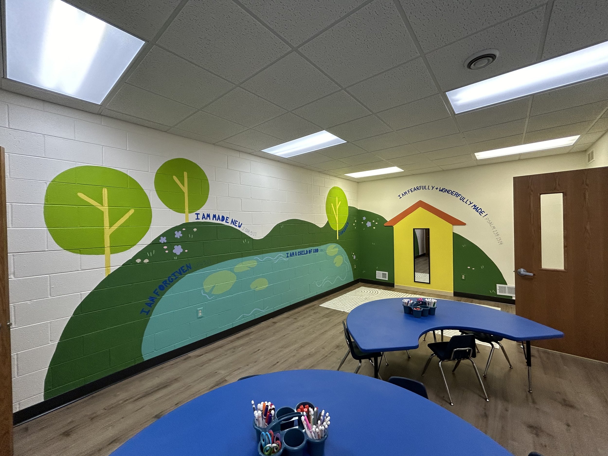 Colorful mural on classroom wall with trees, house, and motivational words like 'I am fearfully and wonderfully made' and 'I am a child of God' in a school classroom with blue tables and chairs.