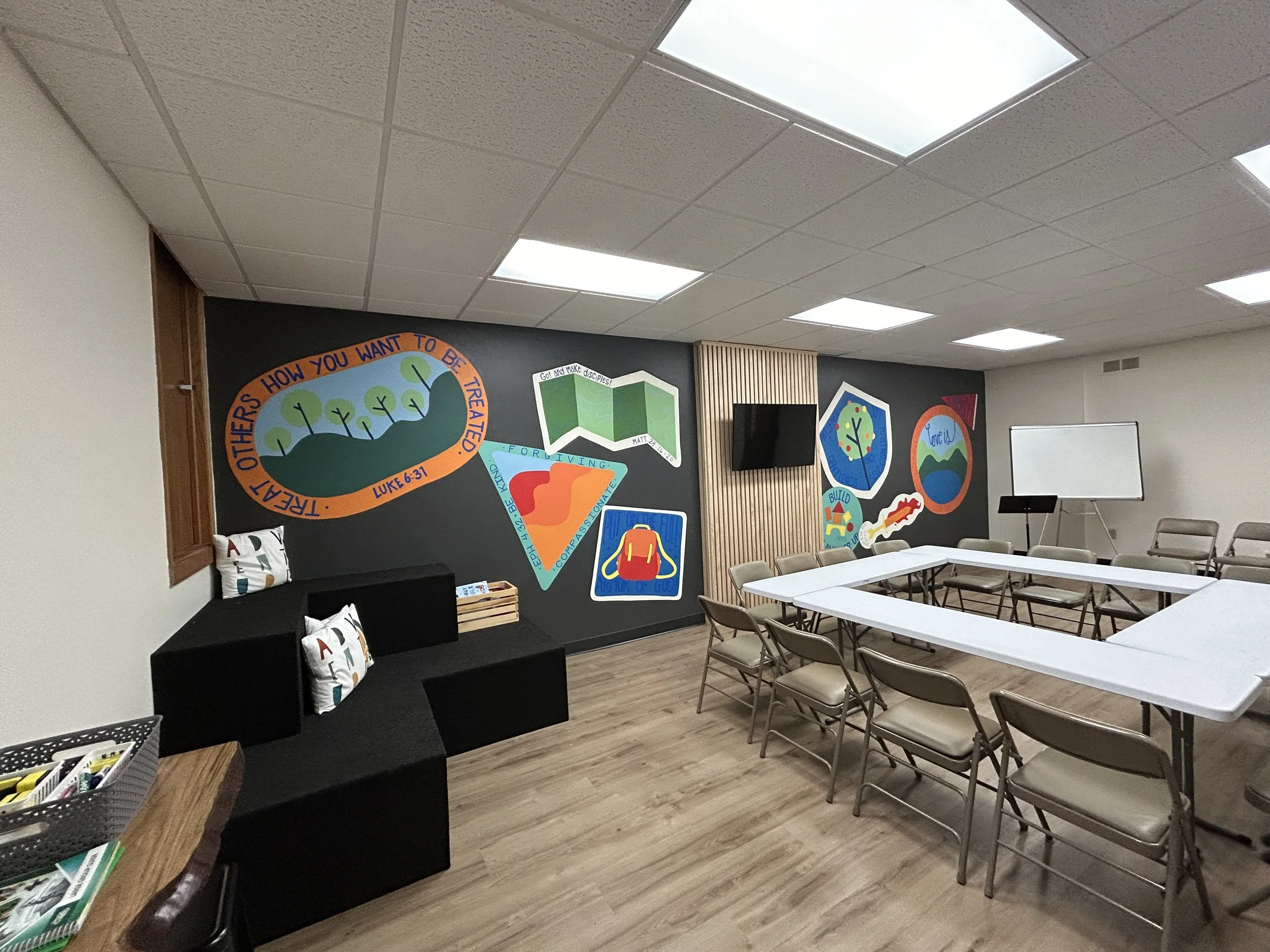 A classroom or meeting room with tables and chairs, decorated with colorful educational posters and a television mounted on a wall.