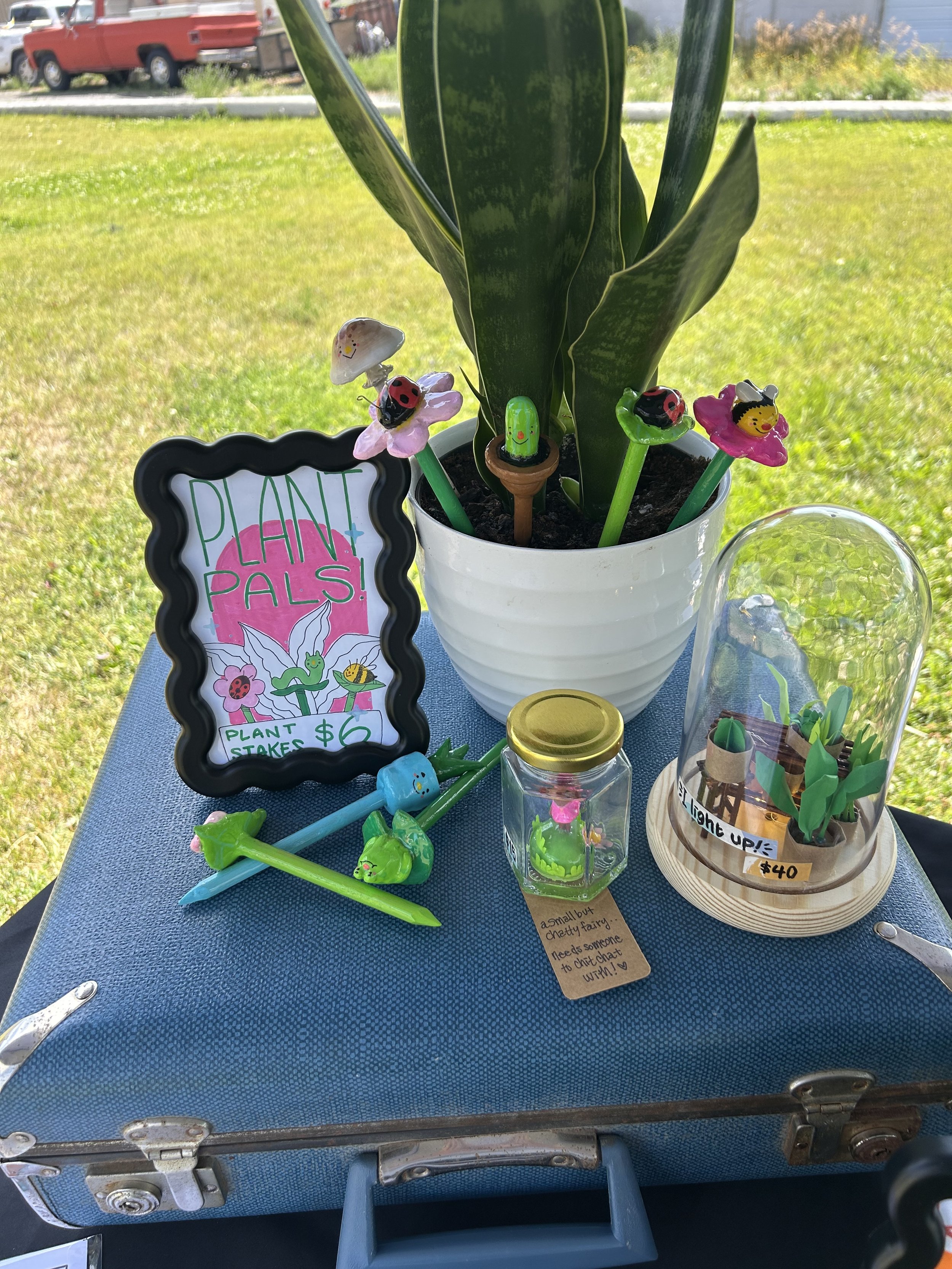 A small table with a blue suitcase on top, decorated with colorful plant-themed items, including a sign that says 'Plant Pals!' and small figurines of insects and plants. There is a white flower pot with a green leafy plant, and several small painted plant and insect figurines, including ladybugs, a bee, and a caterpillar. A clear glass jar with a yellow lid contains small toy insects. There is also a decorative glass dome with green paper plants inside. A handwritten note speaks about a fairy and someone to chat with.