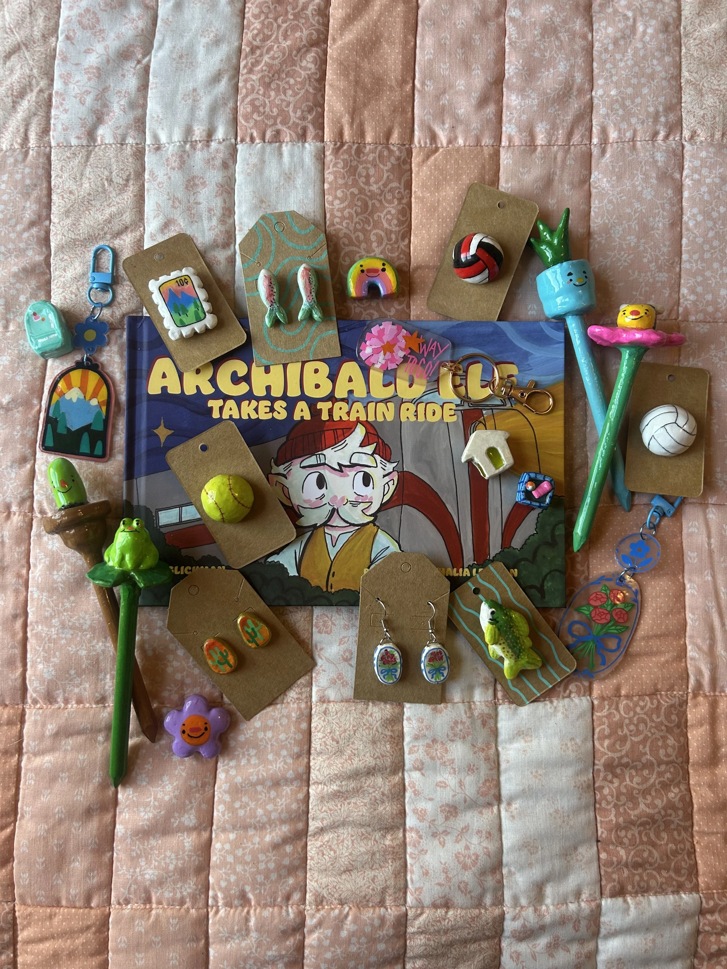Collection of colorful enamel pins, keychains, and jewelry on a pink quilted fabric background. The items include a mountain scene pin, a rainbow figure pin, a volleyball pin, a cactus-shaped pin, and various other whimsical pins and charms.