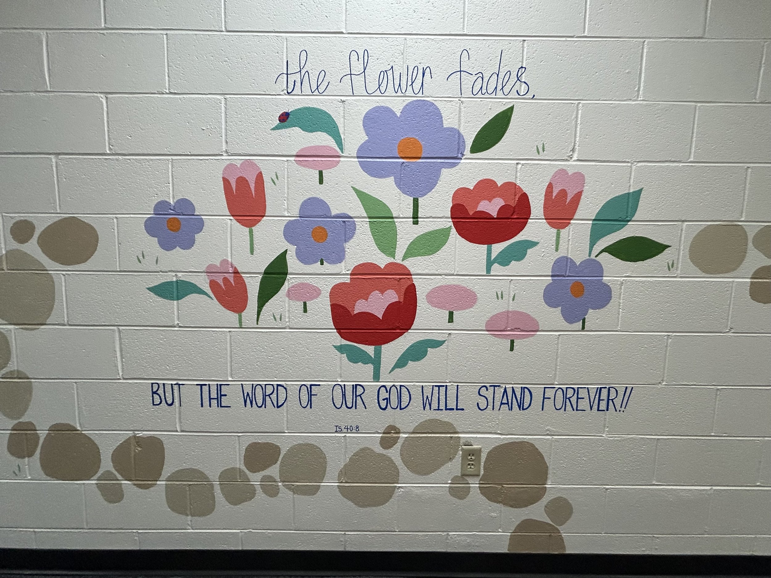 A painted mural on a white brick wall featuring colorful flowers, a bird, and a bible verse from Isaiah 40:8 that reads 'The flower fades but the word of our God will stand forever!'