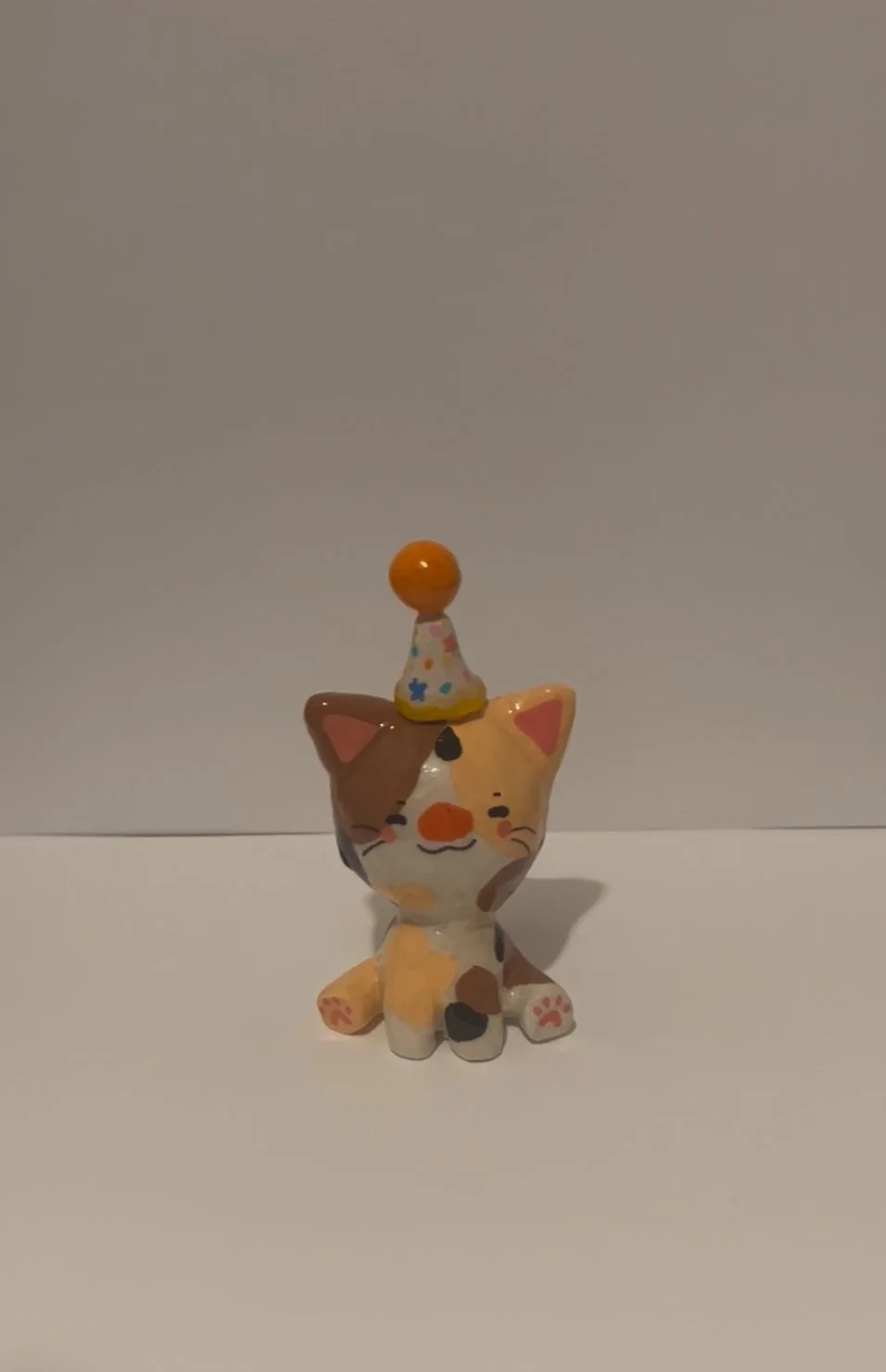 Ceramic figurine of a cute, multicolored, calico cat with closed eyes and a happy expression, wearing a tiny party hat with a pom-pom.