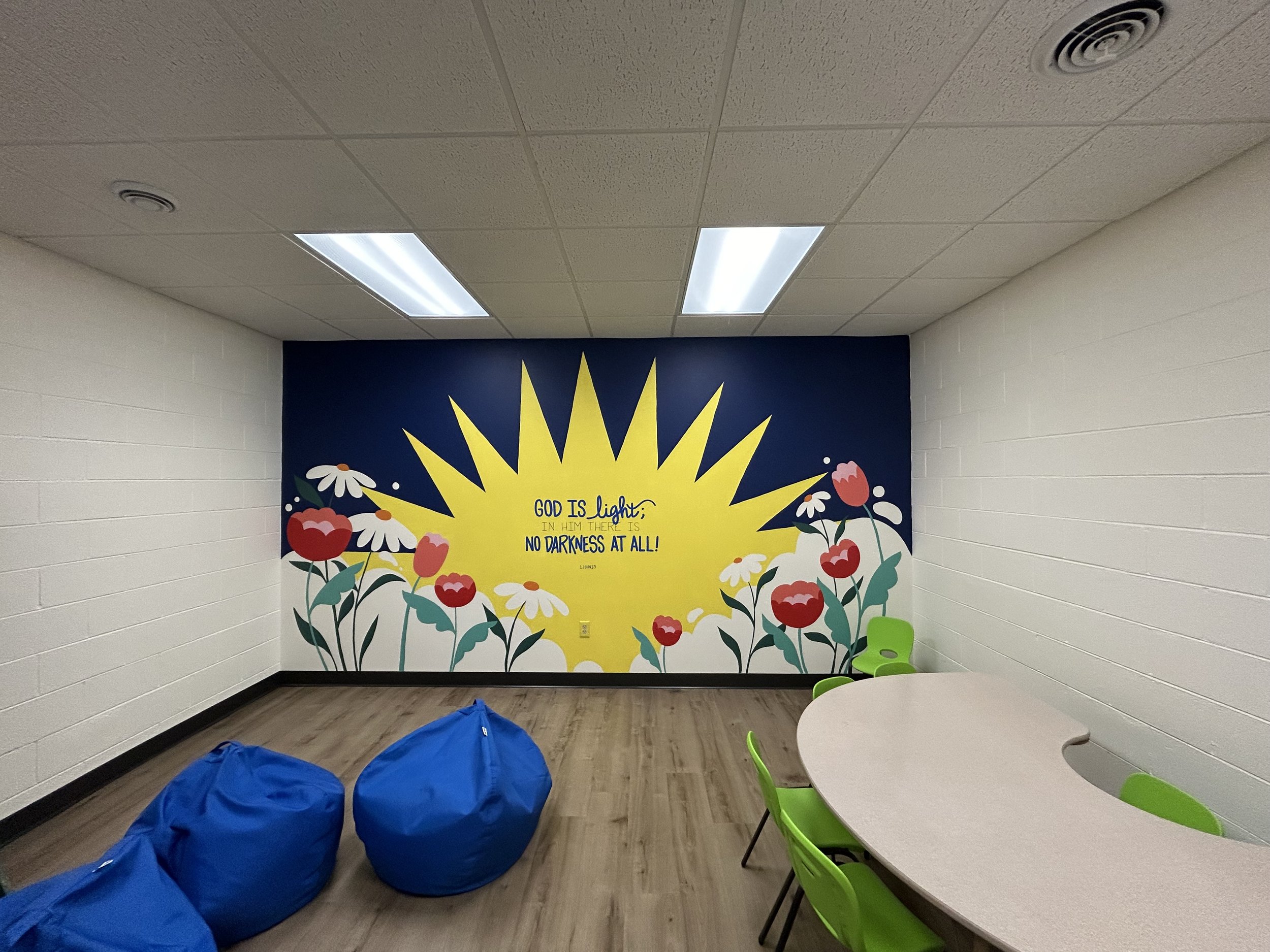A decorated wall with a religious message, surrounded by colorful flowers, in a room with white brick walls and a ceiling with fluorescent lights. Green chairs and blue bean bags are visible.