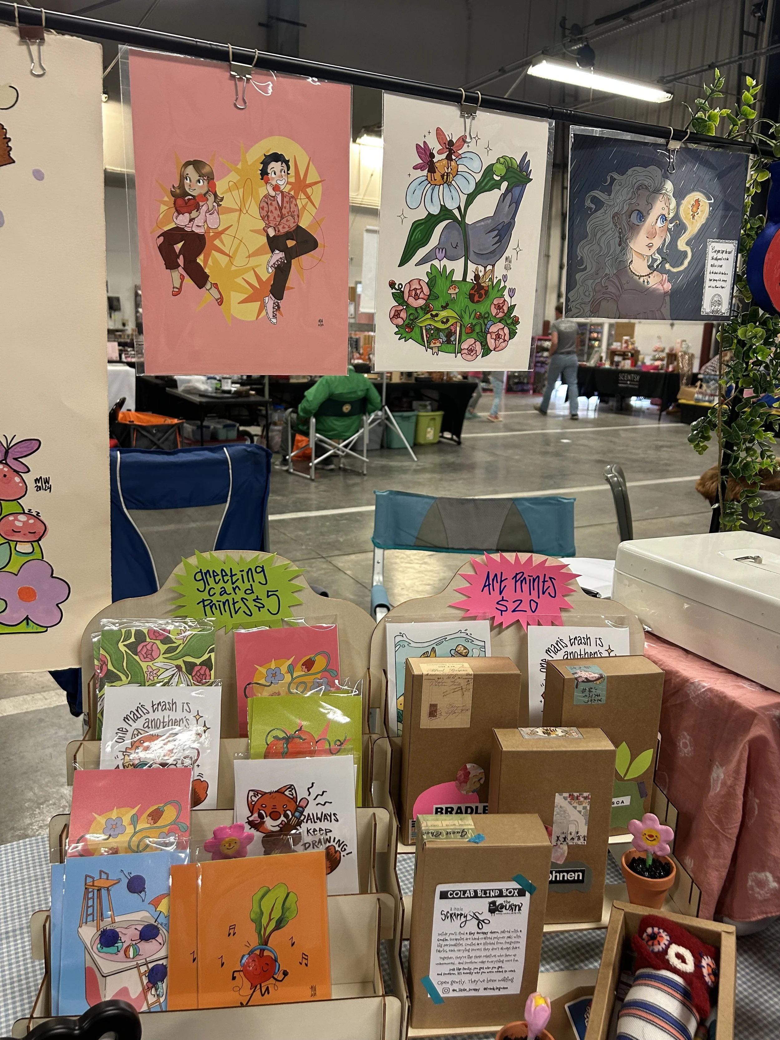 Art prints hanging on display at an indoor market, featuring cartoon-style illustrations of animals and characters with colorful backgrounds and designs.