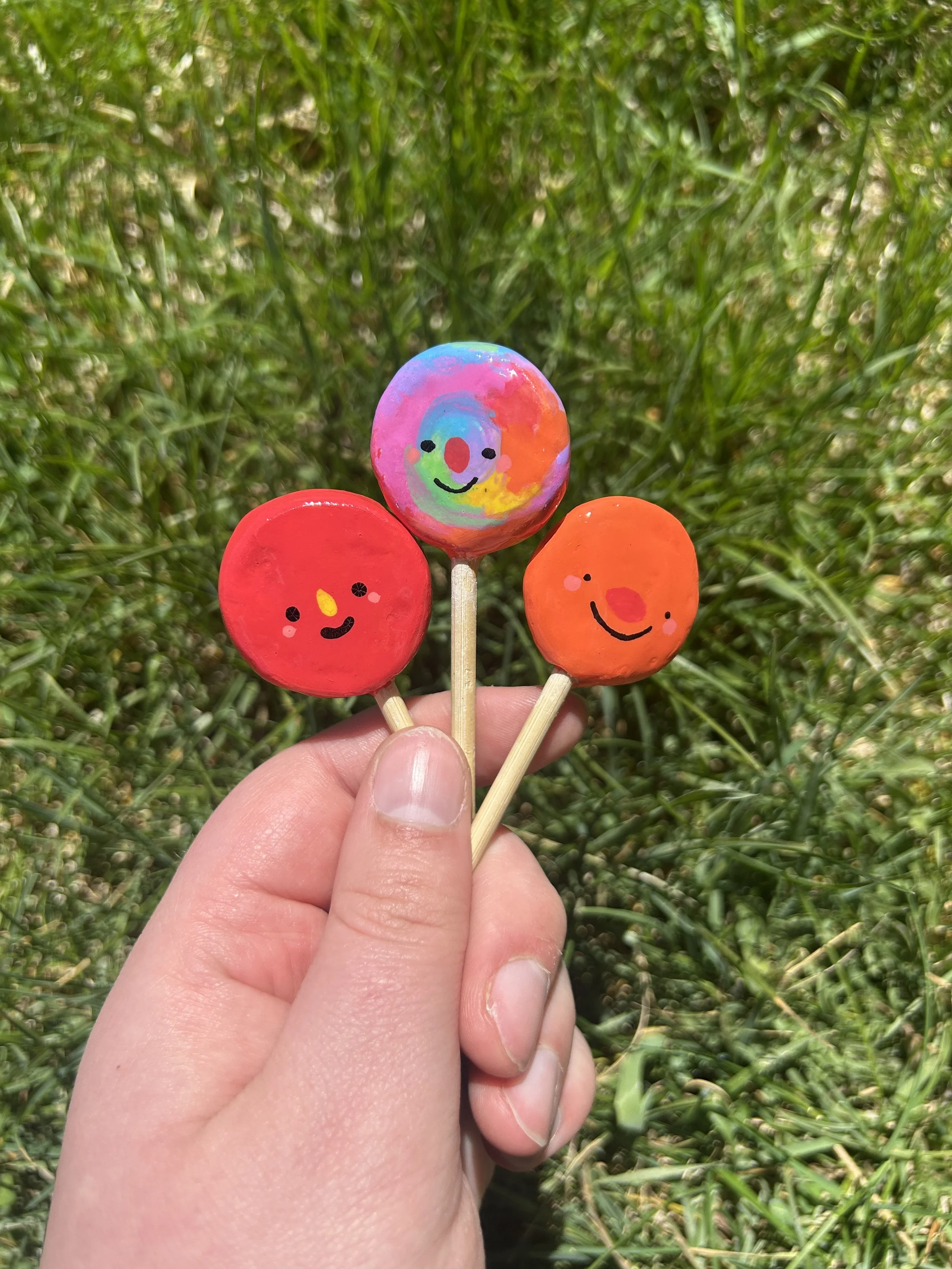 Hand holding three colorful painted lollipops with smiley faces against a background of green grass.