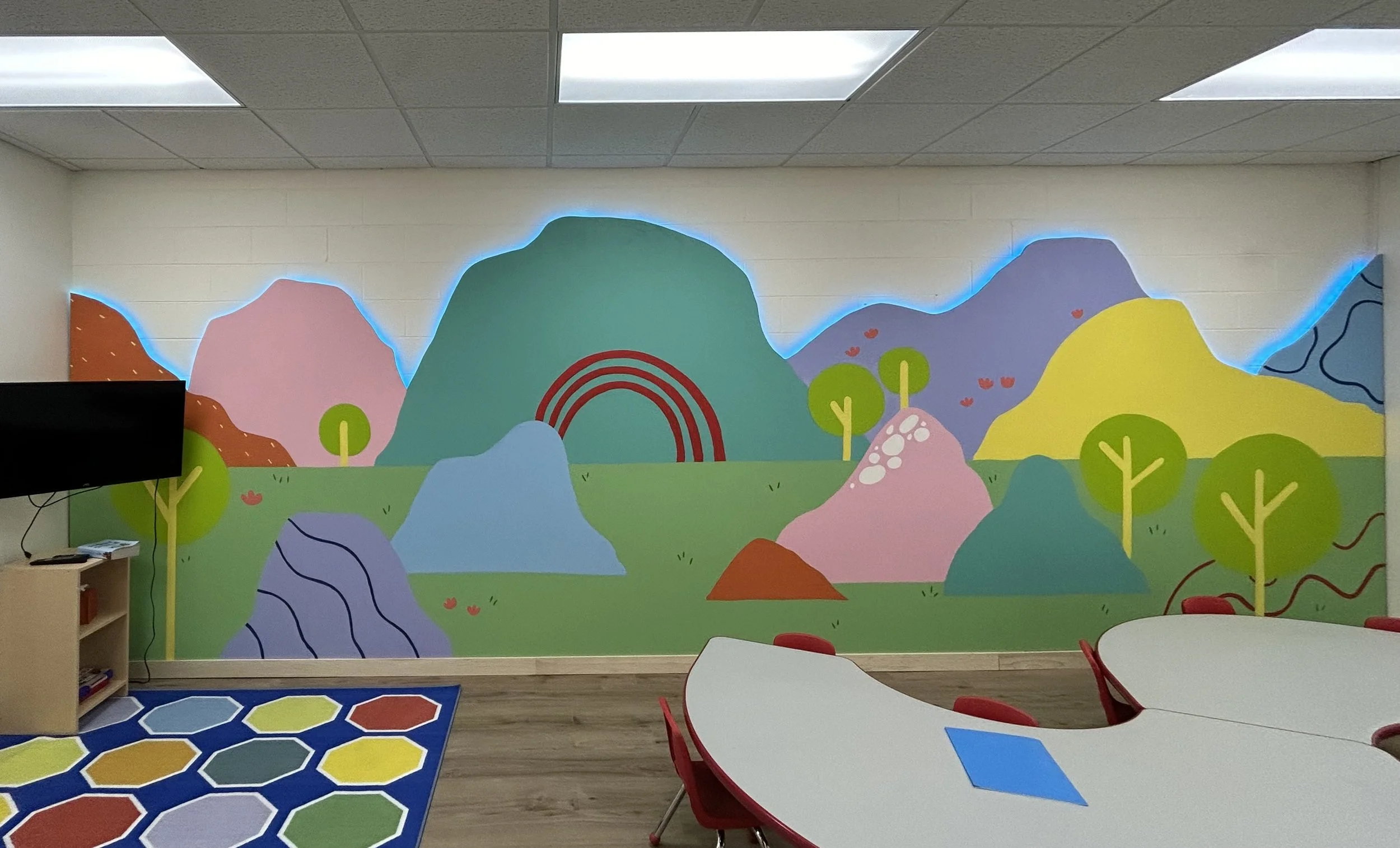 Colorful mural of mountains, trees, and rainbows on a classroom wall, with a blue glow around the painted landscape.