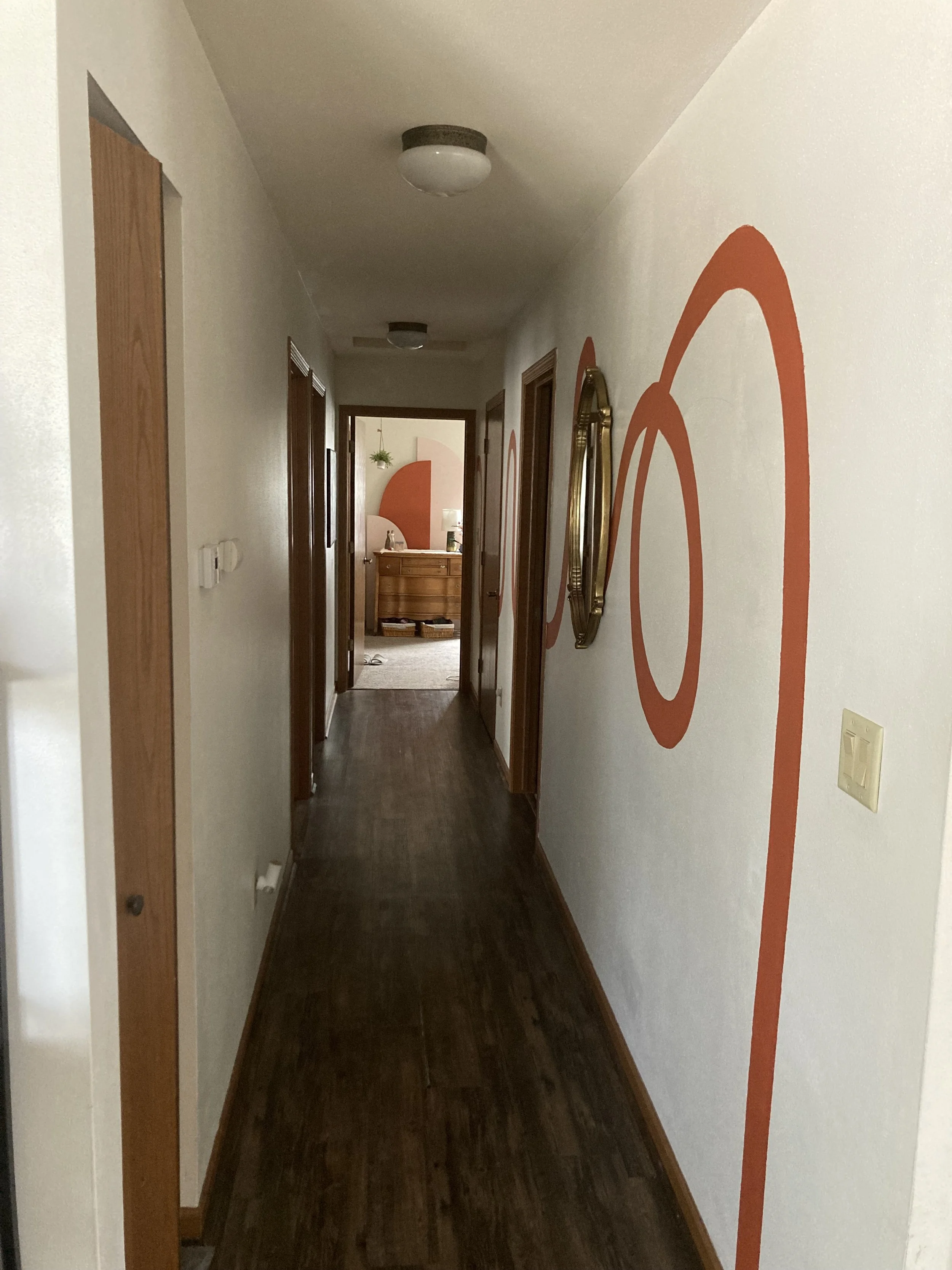 Long hallway with wooden flooring, white walls decorated with orange and pink circular art, and wooden door frames, leading to a room at the end with a hanging plant and a wooden dresser.