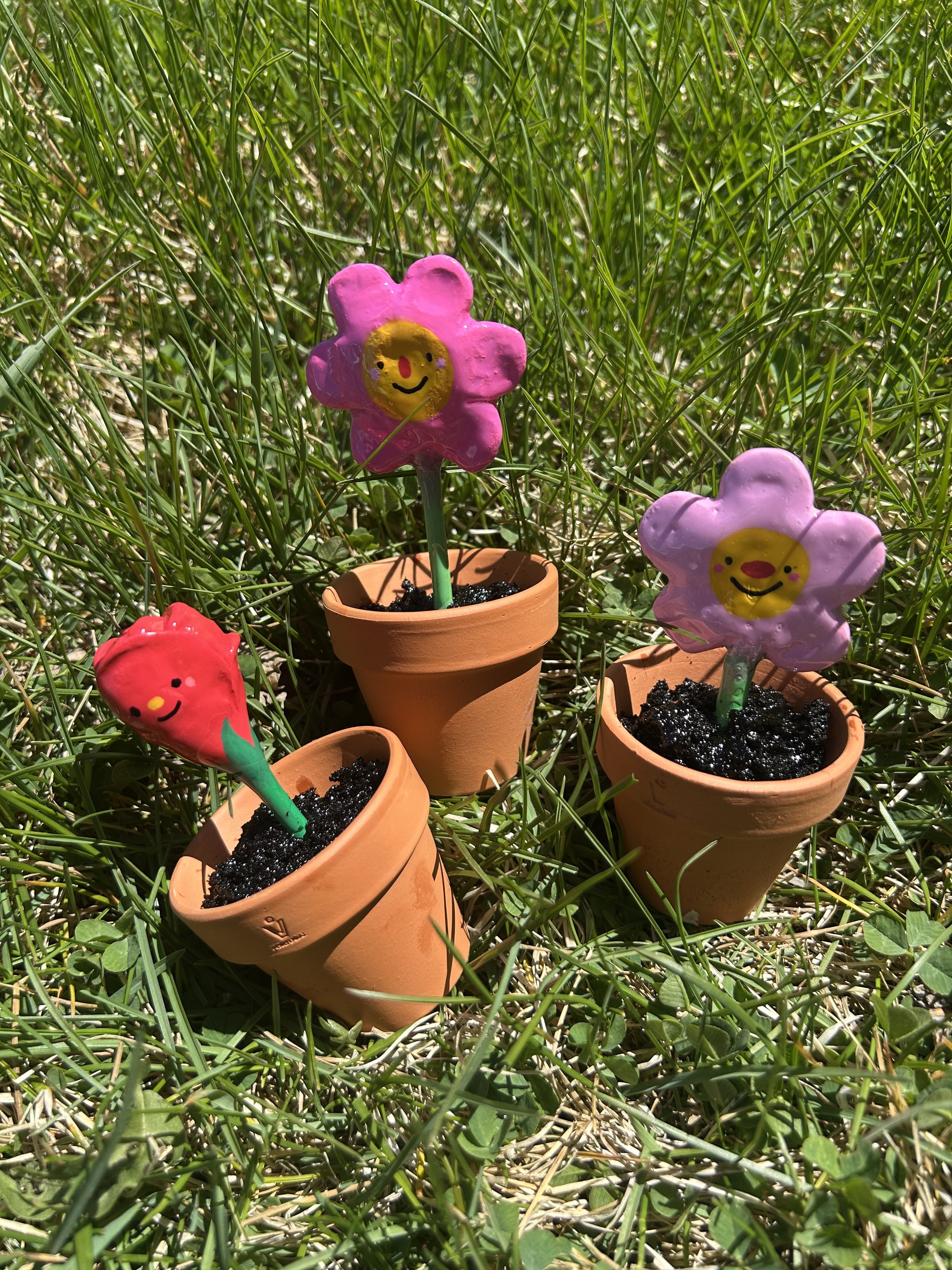 Miniature flower-shaped garden decorations with smiling faces in small terracotta pots on grass.