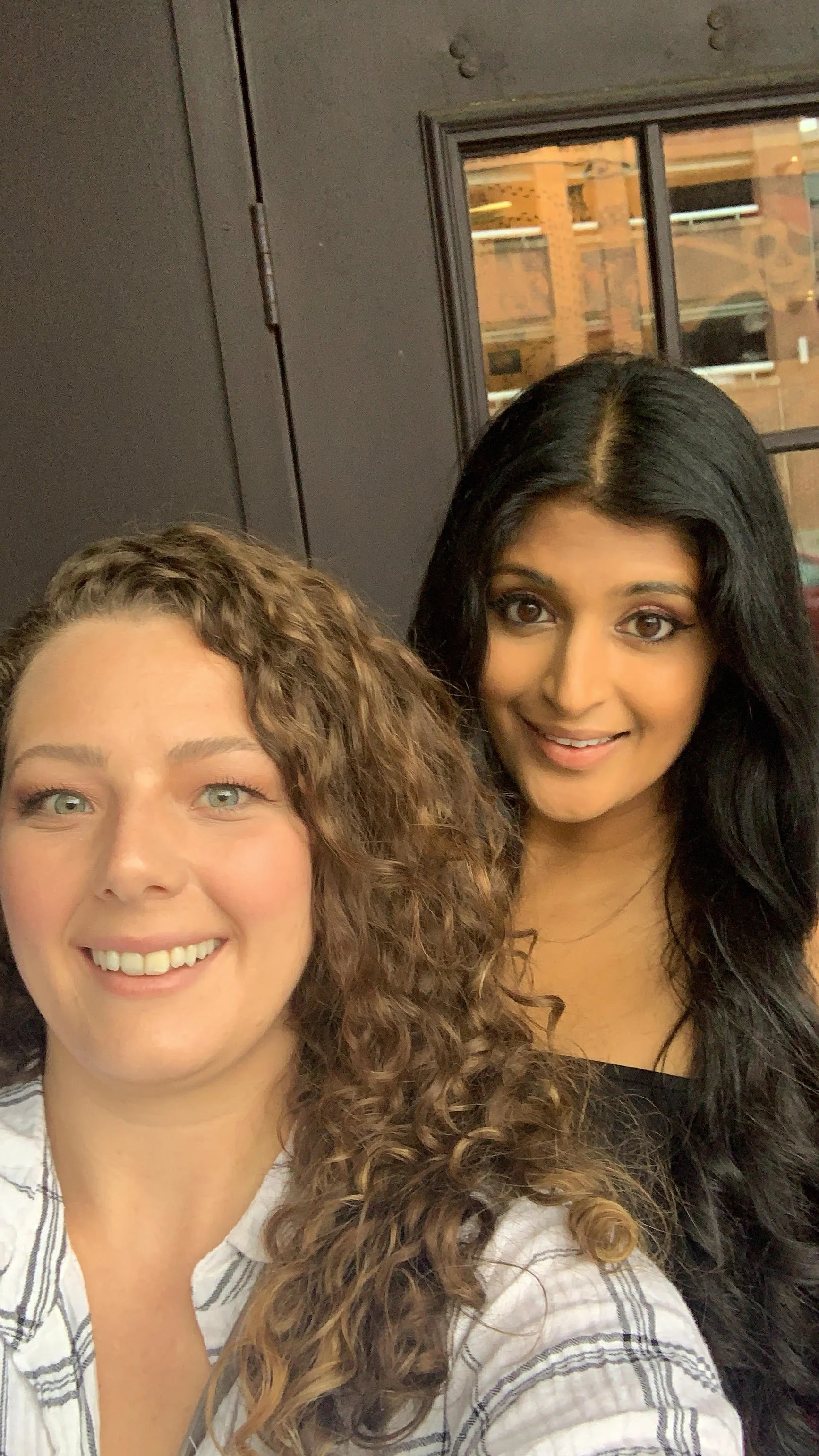 Two women smiling for a selfie indoors, with a window showing a brick building and a street outside.