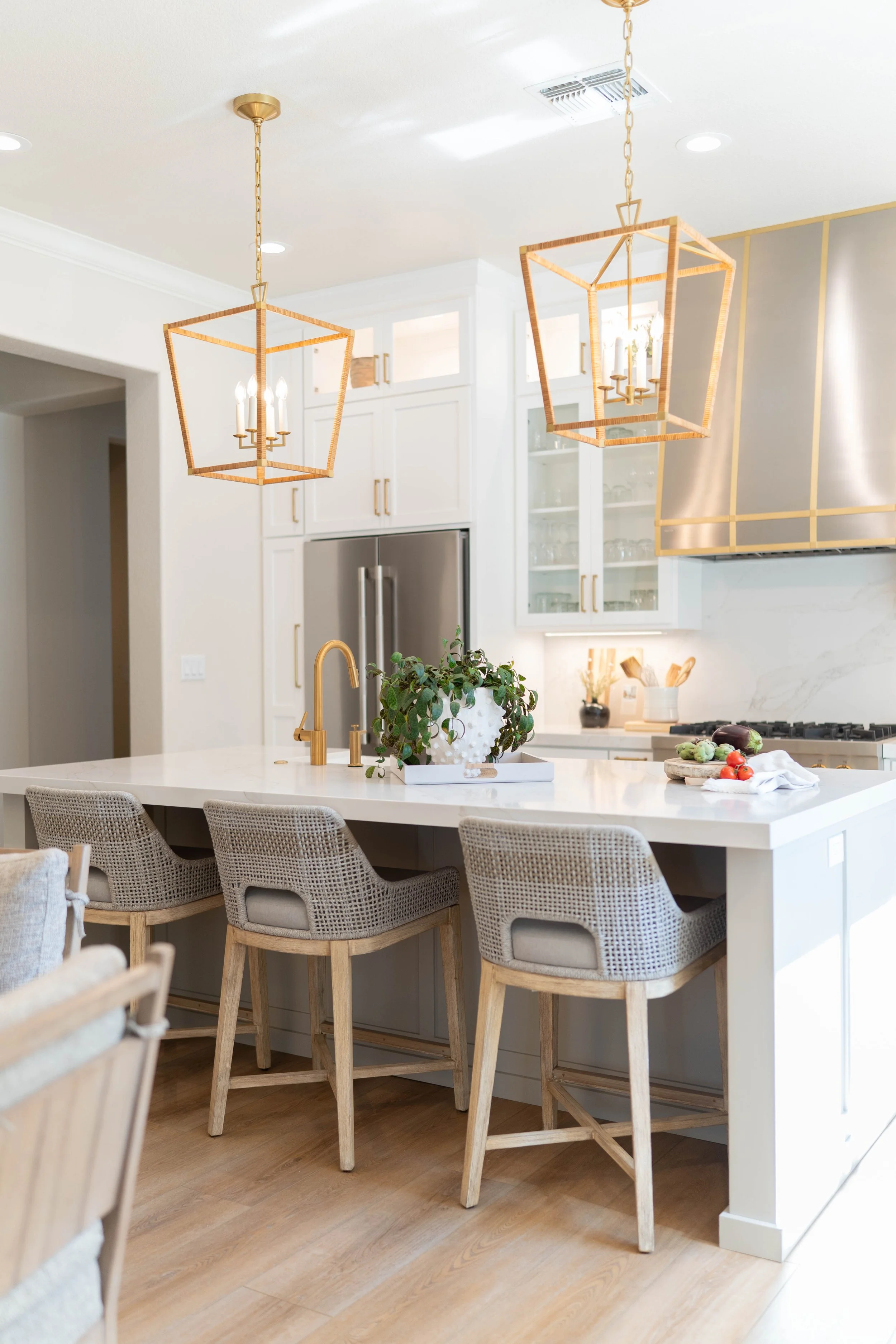 Modern white kitchen with two geometric wooden pendant lights, a kitchen island with beige woven chairs, a gold faucet, a potted plant, and fresh produce on the counter.