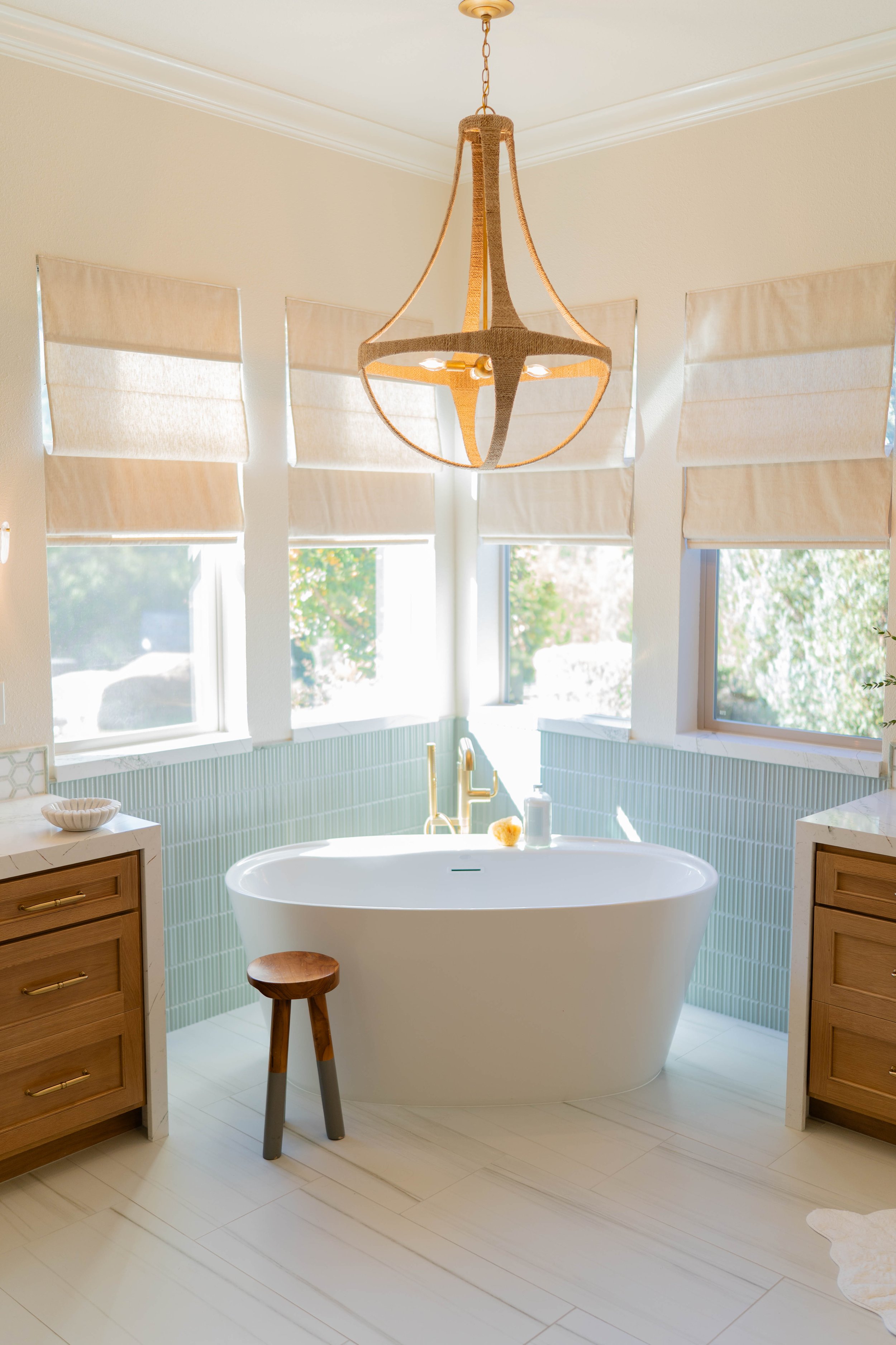 Bright bathroom with a freestanding bathtub near three large windows with beige roman shades, wooden cabinets on either side, and a modern chandelier hanging from the ceiling.
