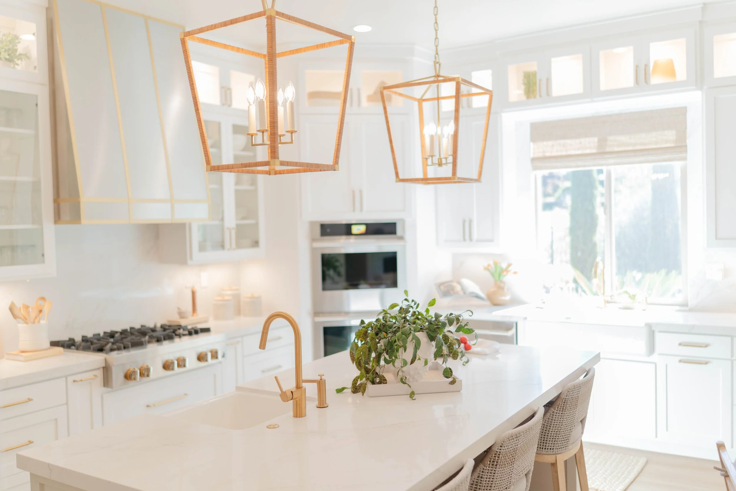 Bright, white kitchen with an island, gold fixtures, modern pendant lights, and large windows letting in natural light.