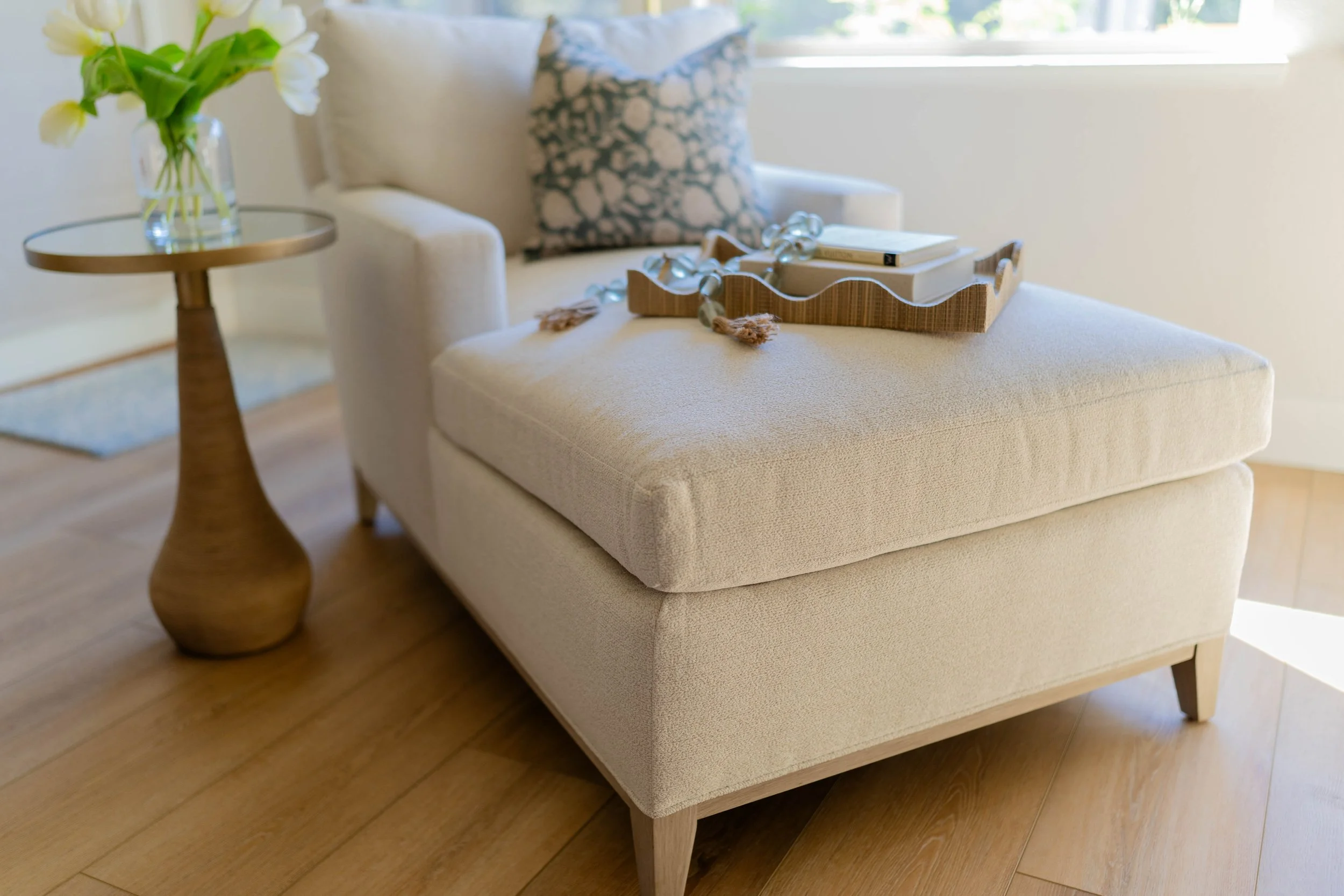 A living room with a cream-colored armchair and ottoman, a small wooden side table with a glass vase of white flowers, and a tray with books and decorative ribbons resting on the ottoman. A decorative pillow with a blue and white floral pattern is on the armchair, and sunlight streams through a window.
