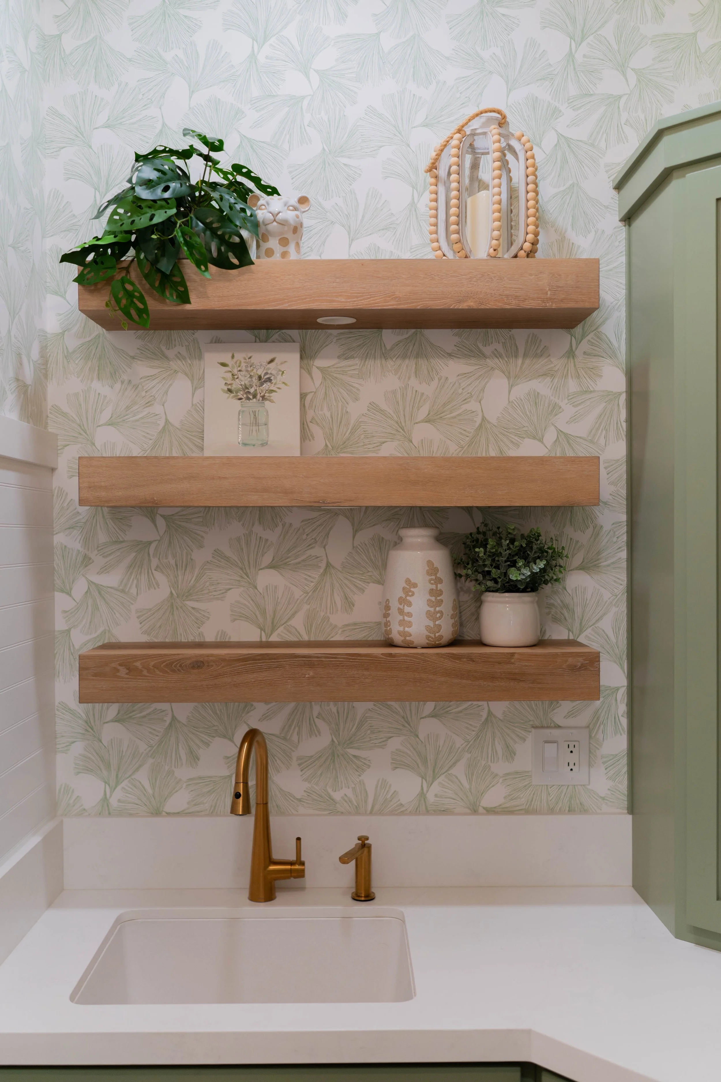 Decorative kitchen area with two wooden floating shelves, patterned wallpaper, a green plant, white vases, and a gold faucet.