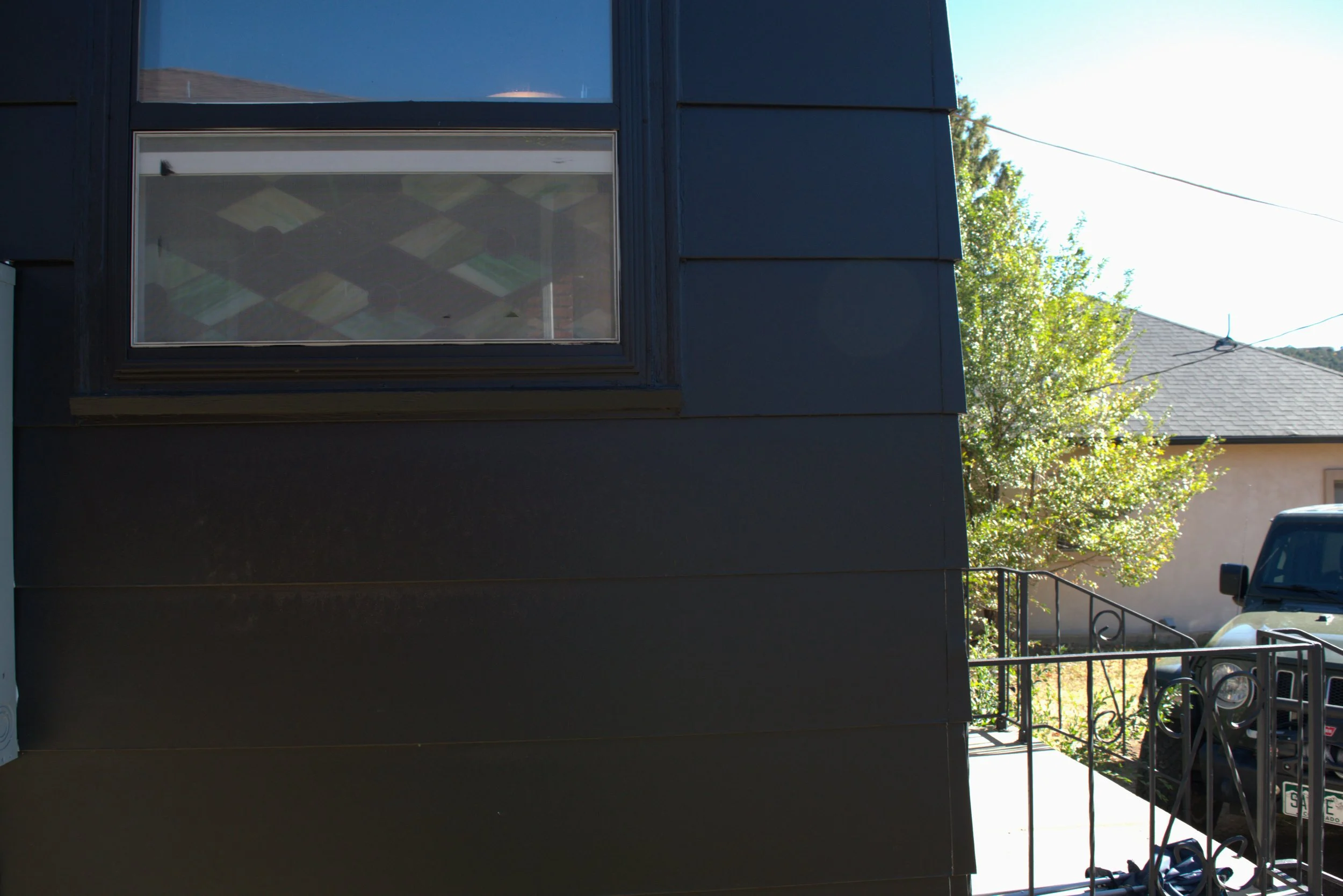 Exterior view of a modern building with black siding, a large window showing an interior ceiling with ceiling tiles, a small front porch with a metal railing, and a parked black vehicle on the driveway with a tree and house visible in the background.