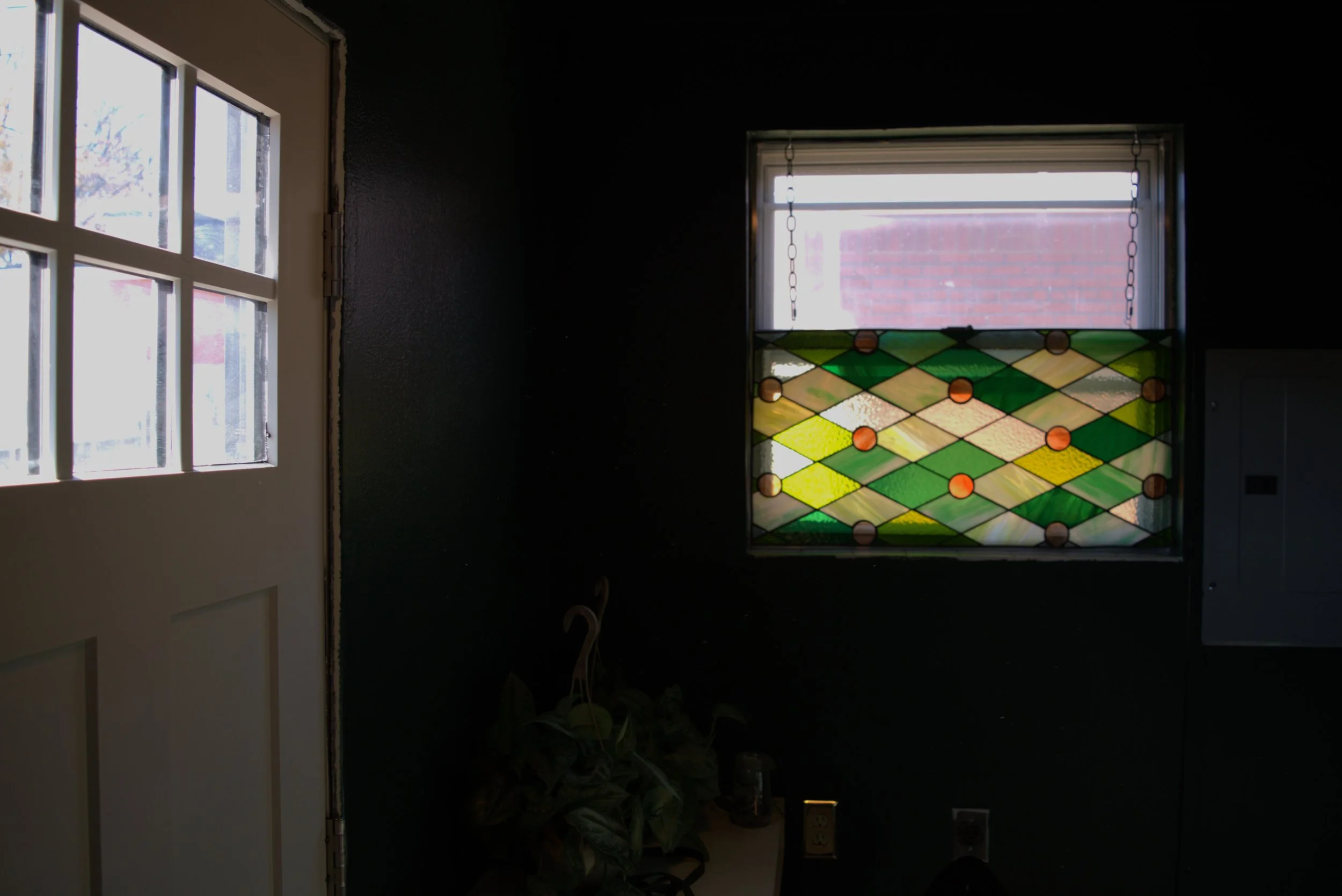 Dark interior room with a stained glass window featuring green, yellow, and orange diamond shapes, and a small window above it. A potted plant and electrical outlets are visible near the bottom.