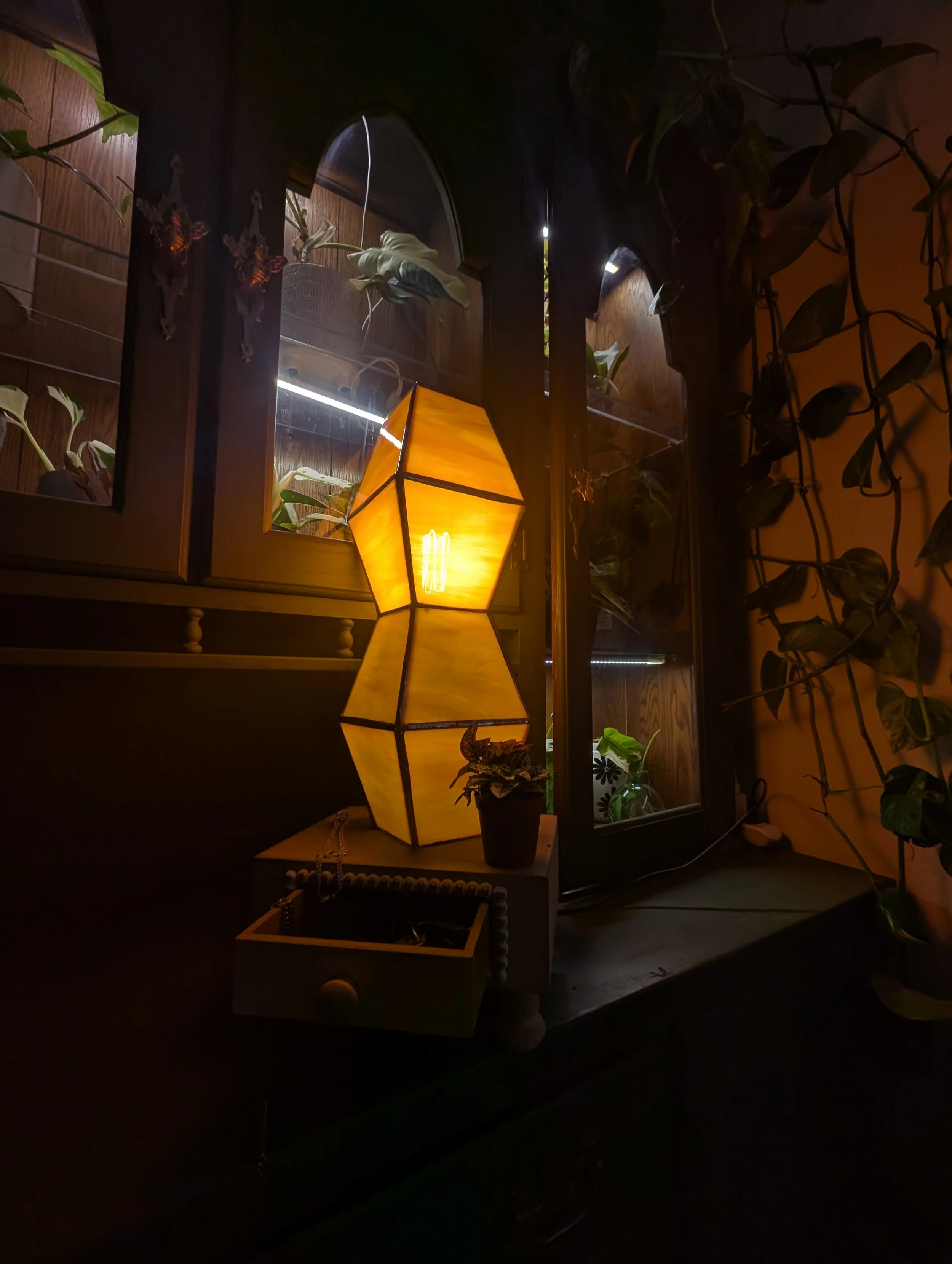 A warm yellow geometric lamp on a dark wooden surface, with potted plants nearby, against a dark background with a window and hanging plants.