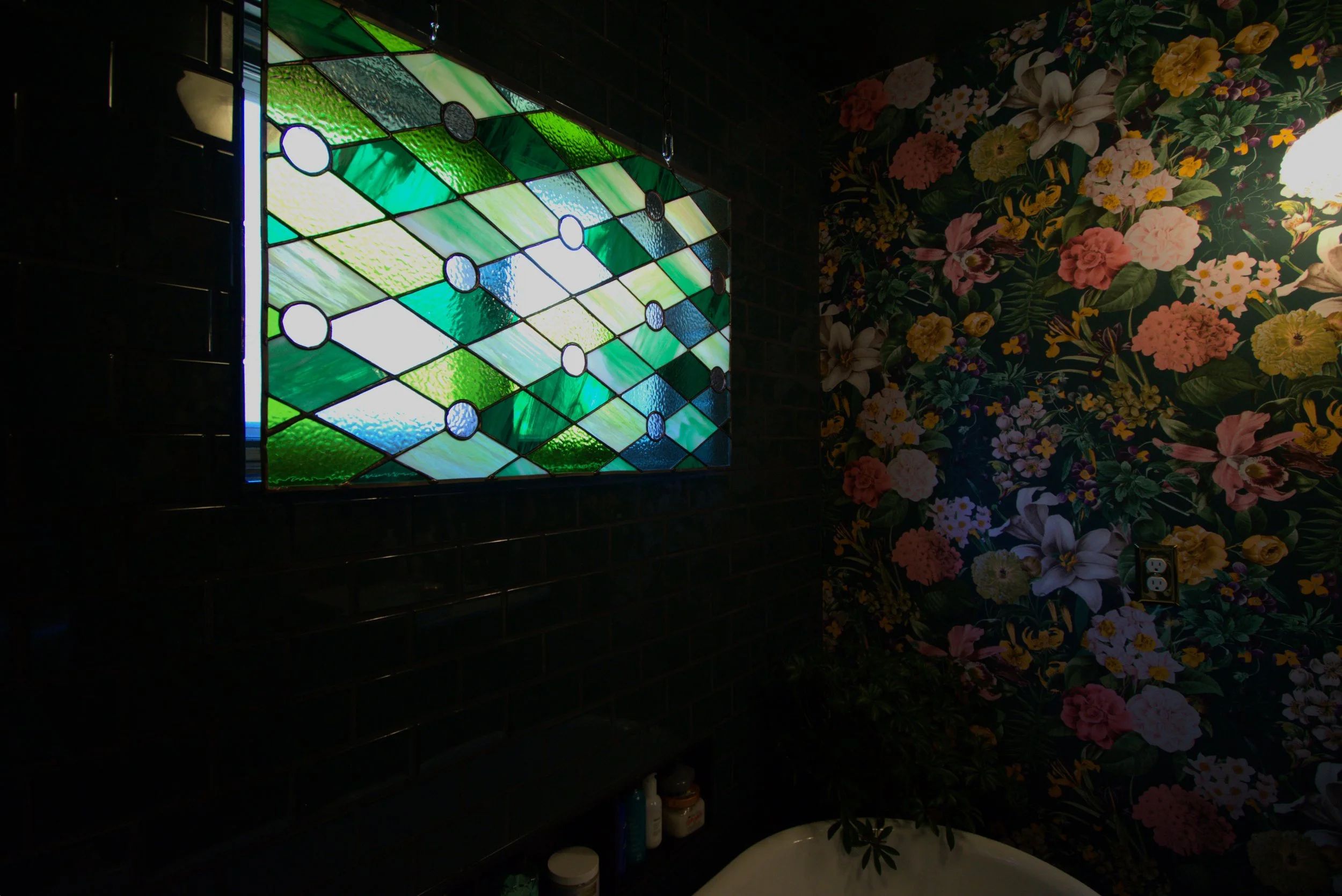 A colorful stained glass window with green and blue geometric patterns, next to a floral wallpaper with large pink, white, and yellow flowers, and a small section of a bathtub.