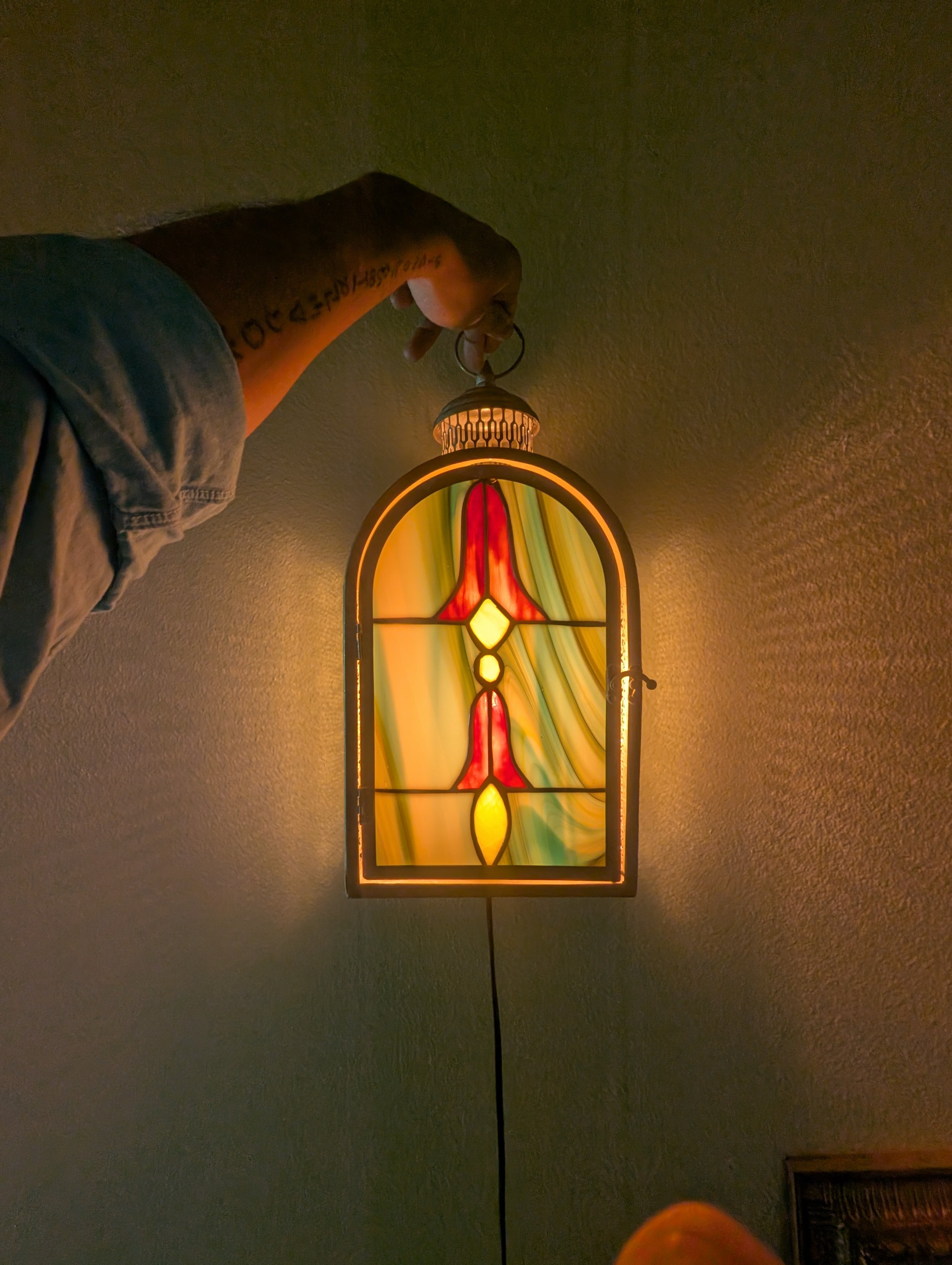 Person holding a stained glass lamp with colorful design, standing or hanging against a beige wall.