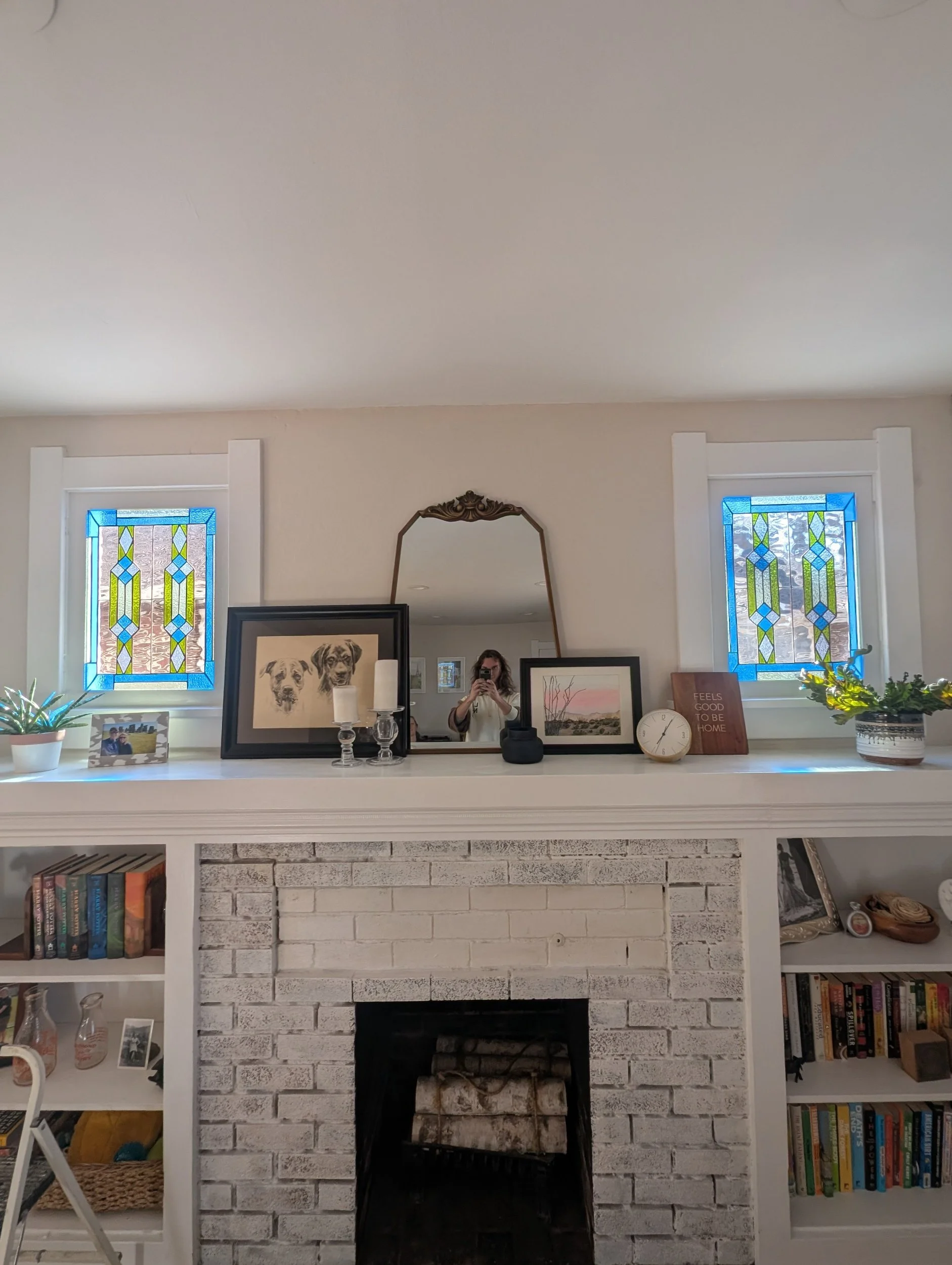 Living room fireplace with white brick, black interior, and white mantel decorated with framed artwork, lamps, candles, clock, and plants, flanked by two stained glass windows.