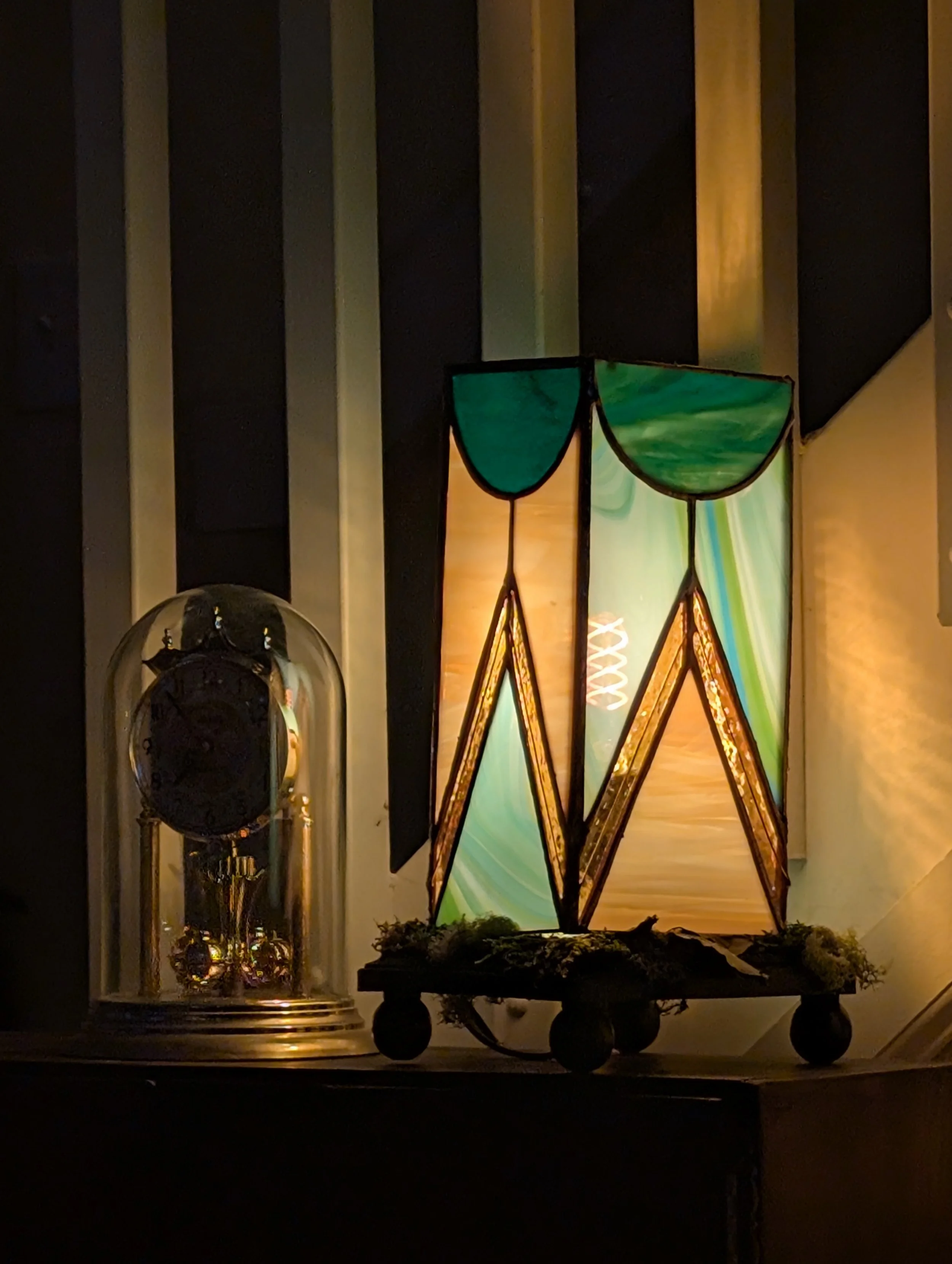 Decorative lamp with green, blue, and orange hues on a wooden surface next to a glass-domed clock with gold accents.