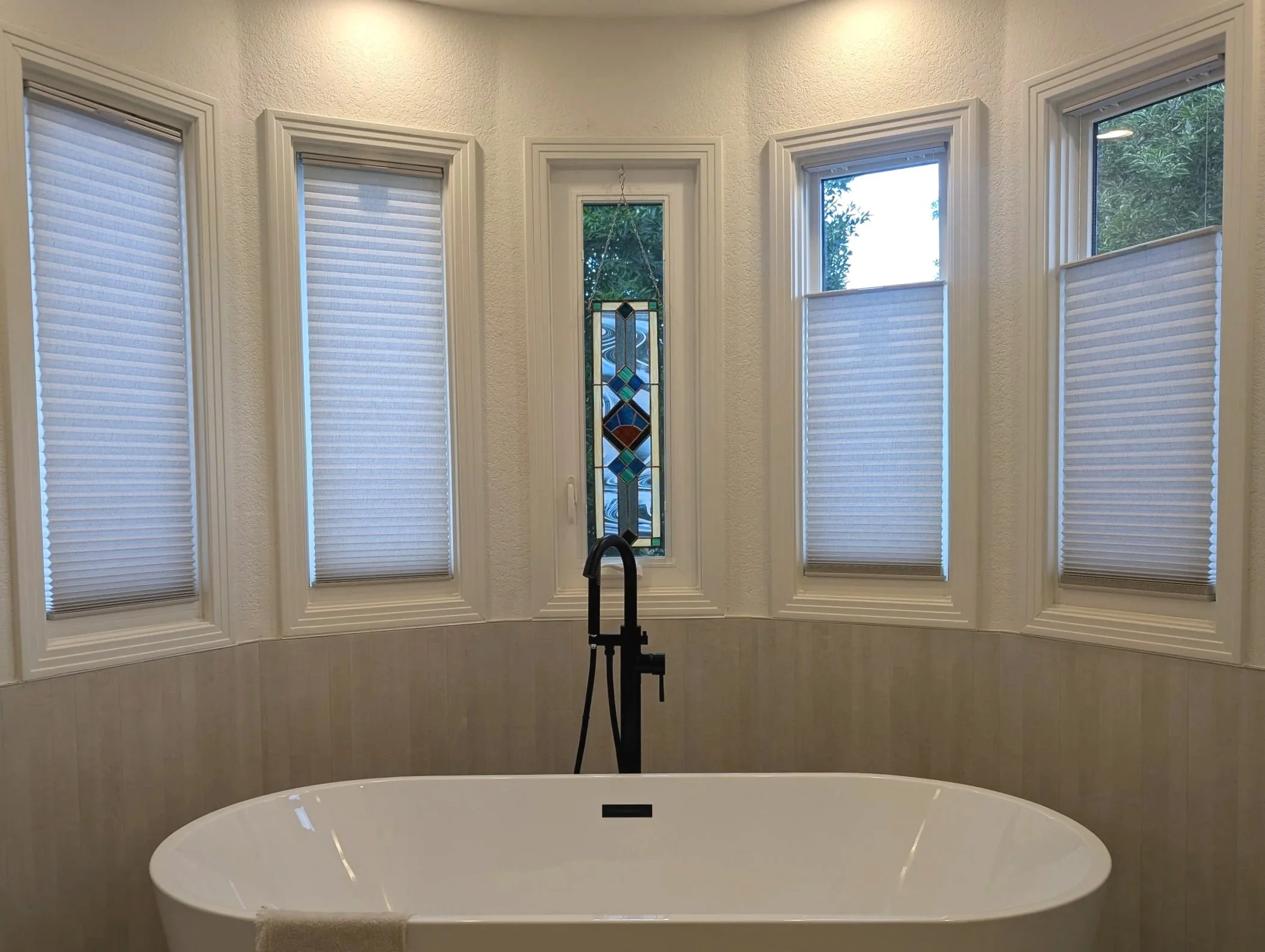 A bathroom with a freestanding bathtub, five windows with white blinds, a decorative stained glass window at the center, and a black faucet.