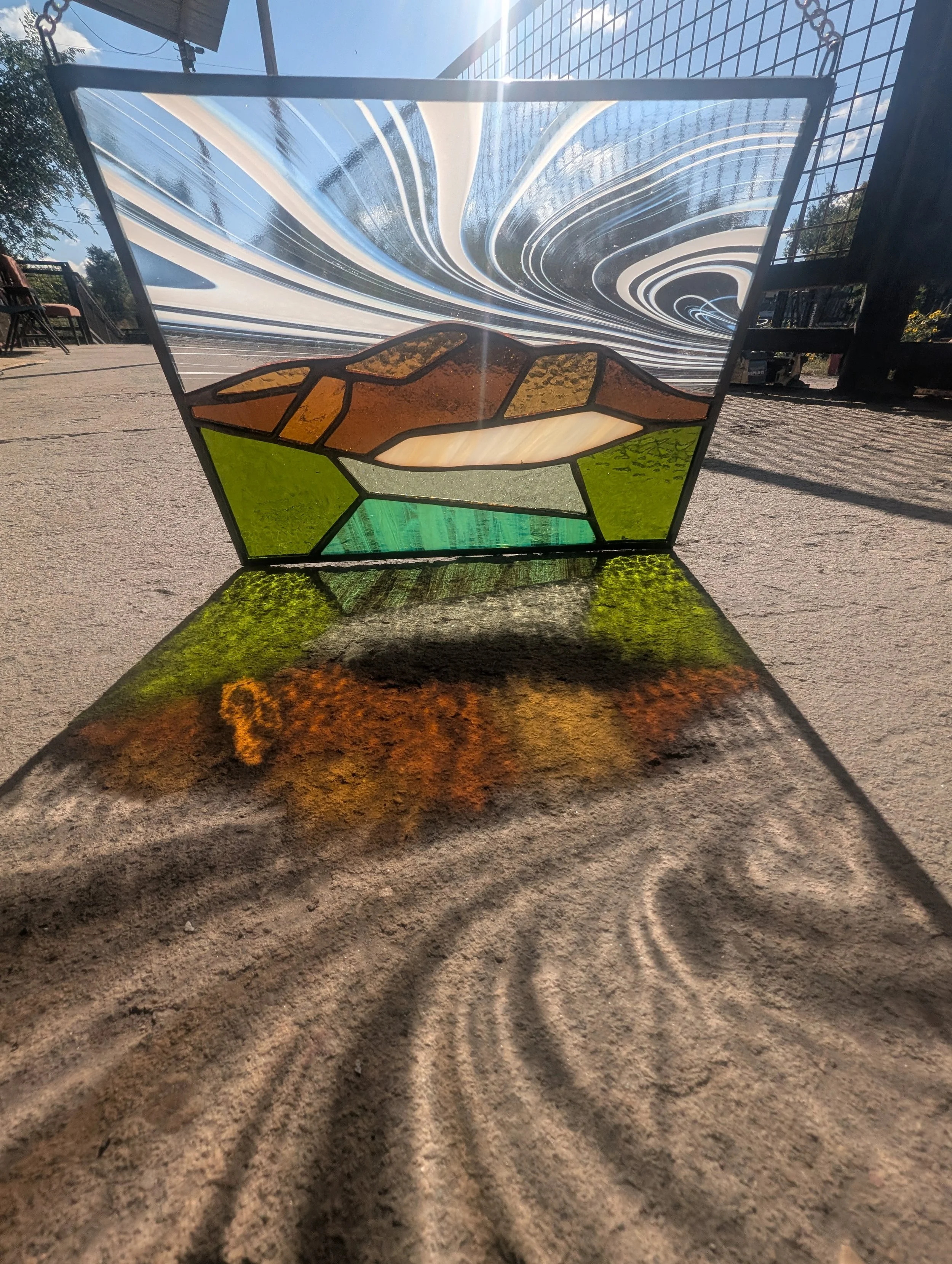 A stained glass panel with a landscape scene featuring mountains, green fields, and a sky with swirling clouds, casting colorful shadows on the ground.
