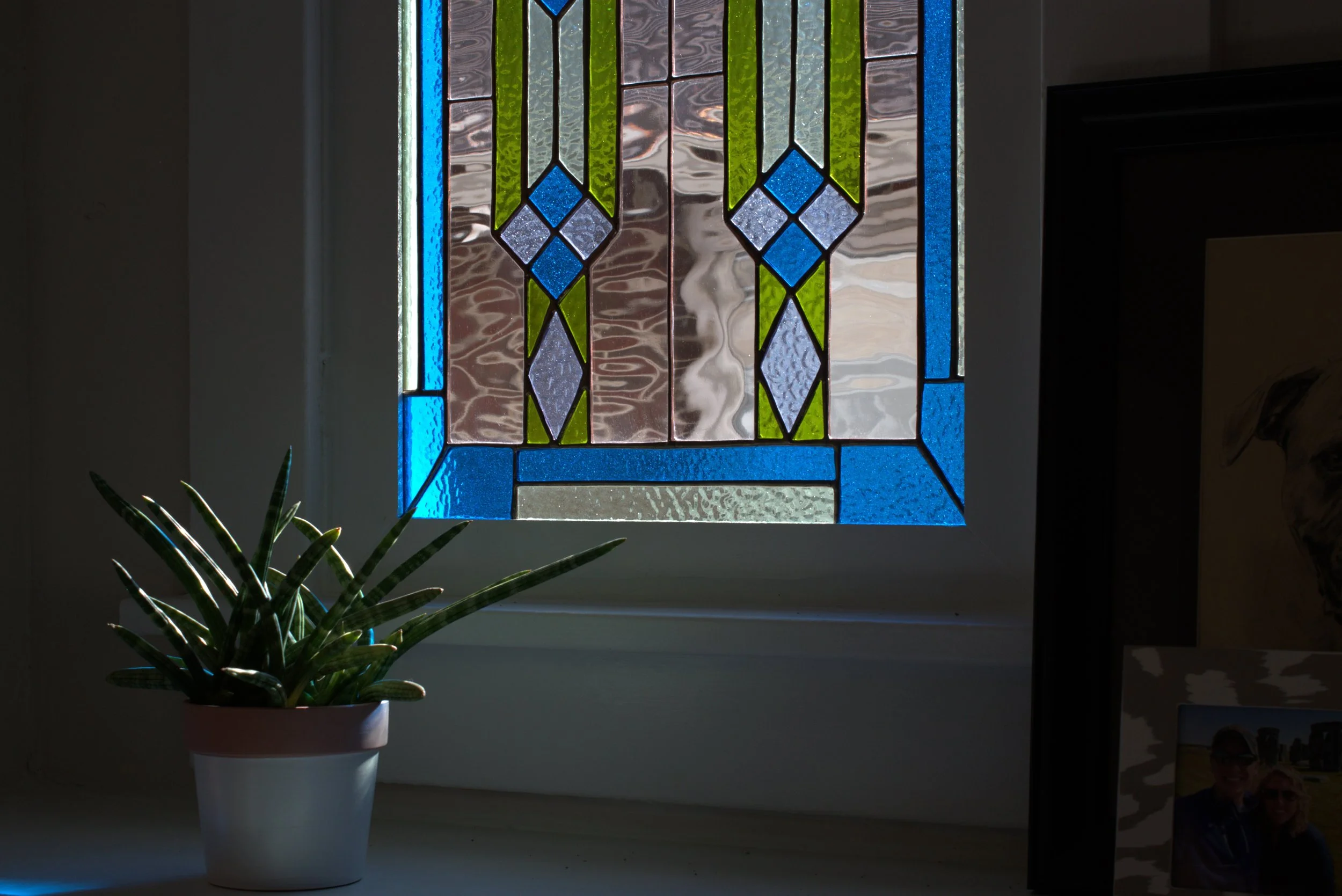Colorful stained glass window with blue, green, and gray geometric patterns, a potted succulent plant in front, and a framed photograph partially visible to the right.