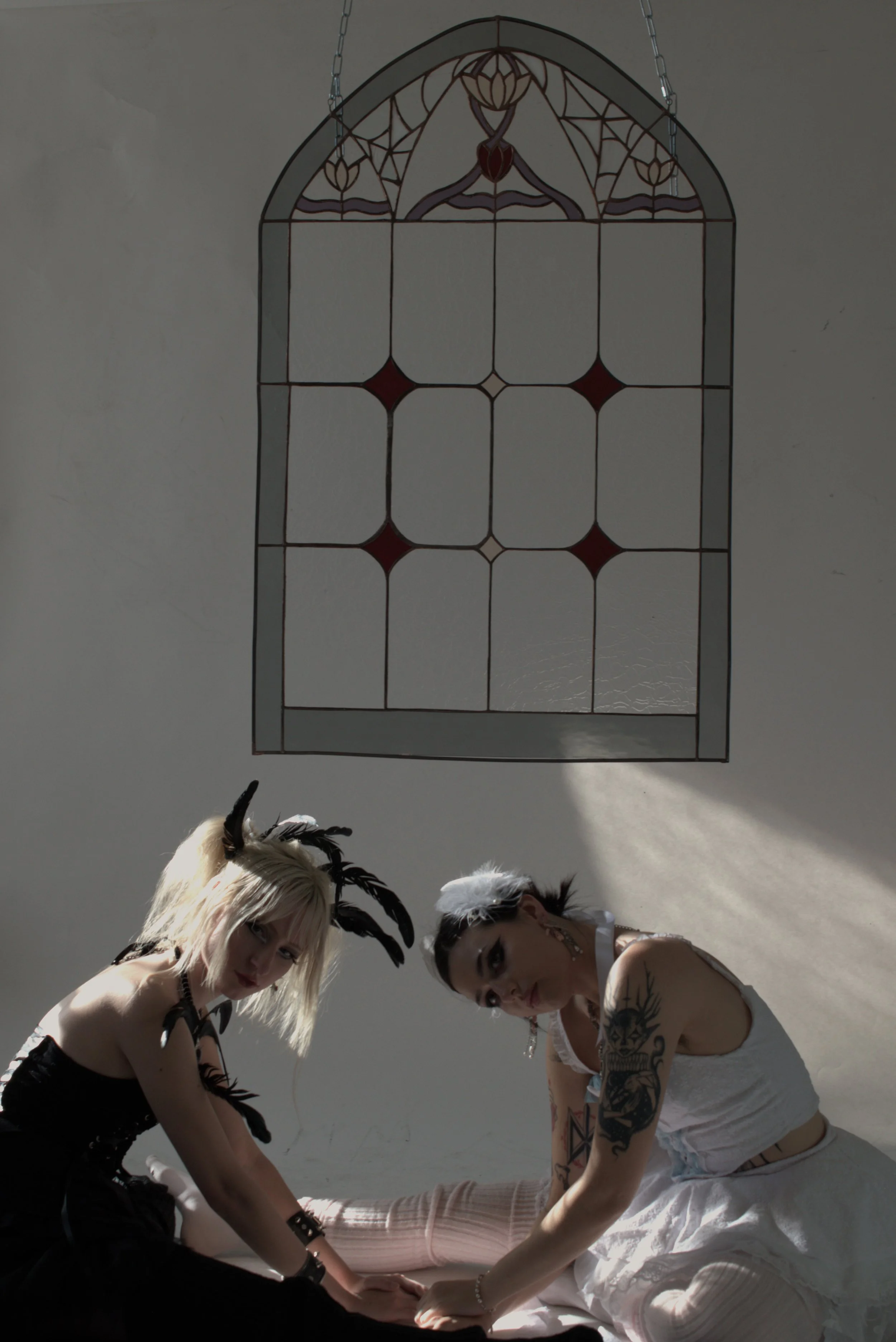 Two women with tattoos and fantasy-themed outfits kneeling on the floor in front of a stained glass window, with dramatic lighting casting shadows.