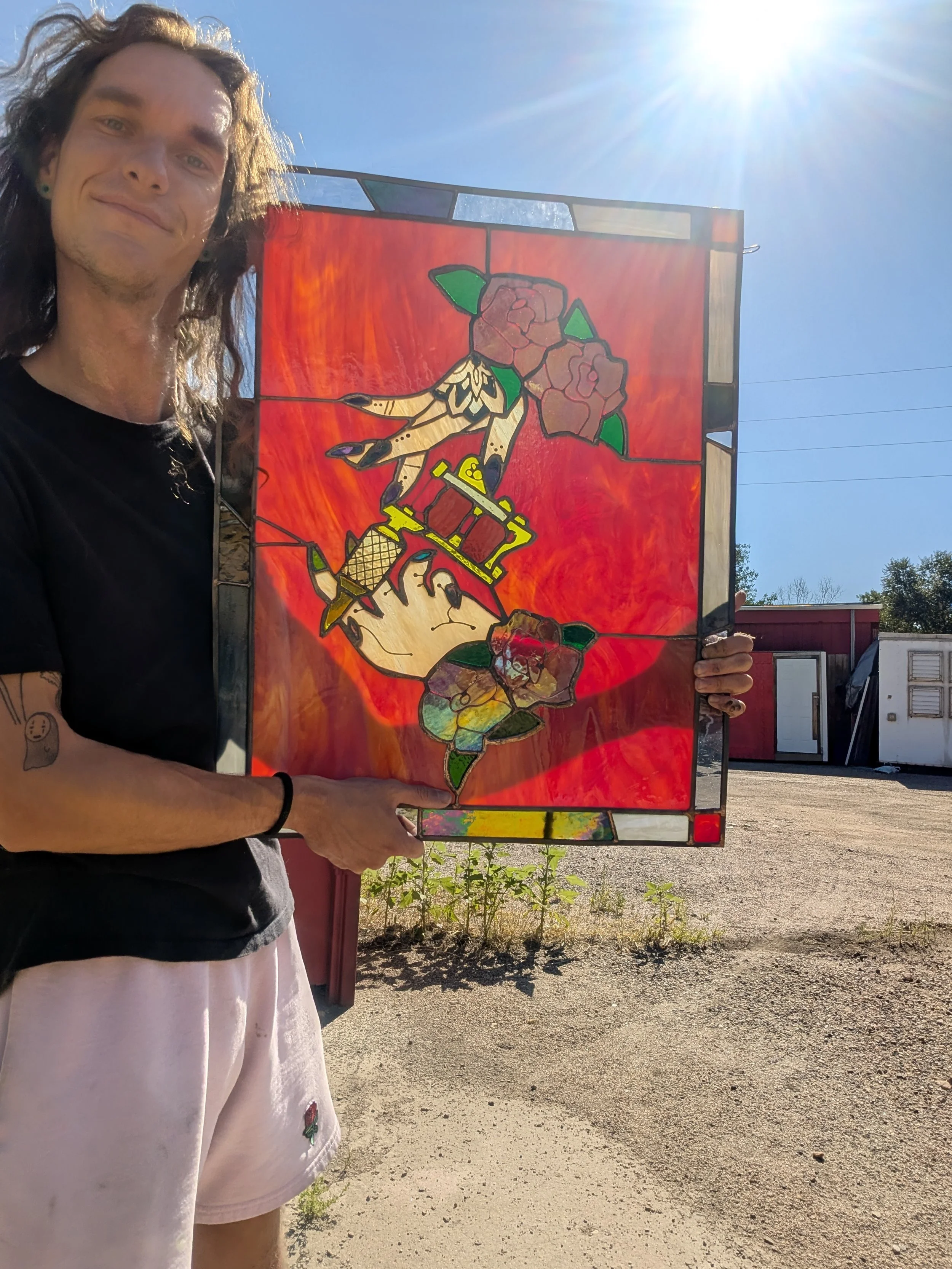 A man with long hair, wearing a black shirt and pink shorts, is holding up a stained glass panel with a rose design. He is outdoors in sunlight, with a red building and some small plants in the background.