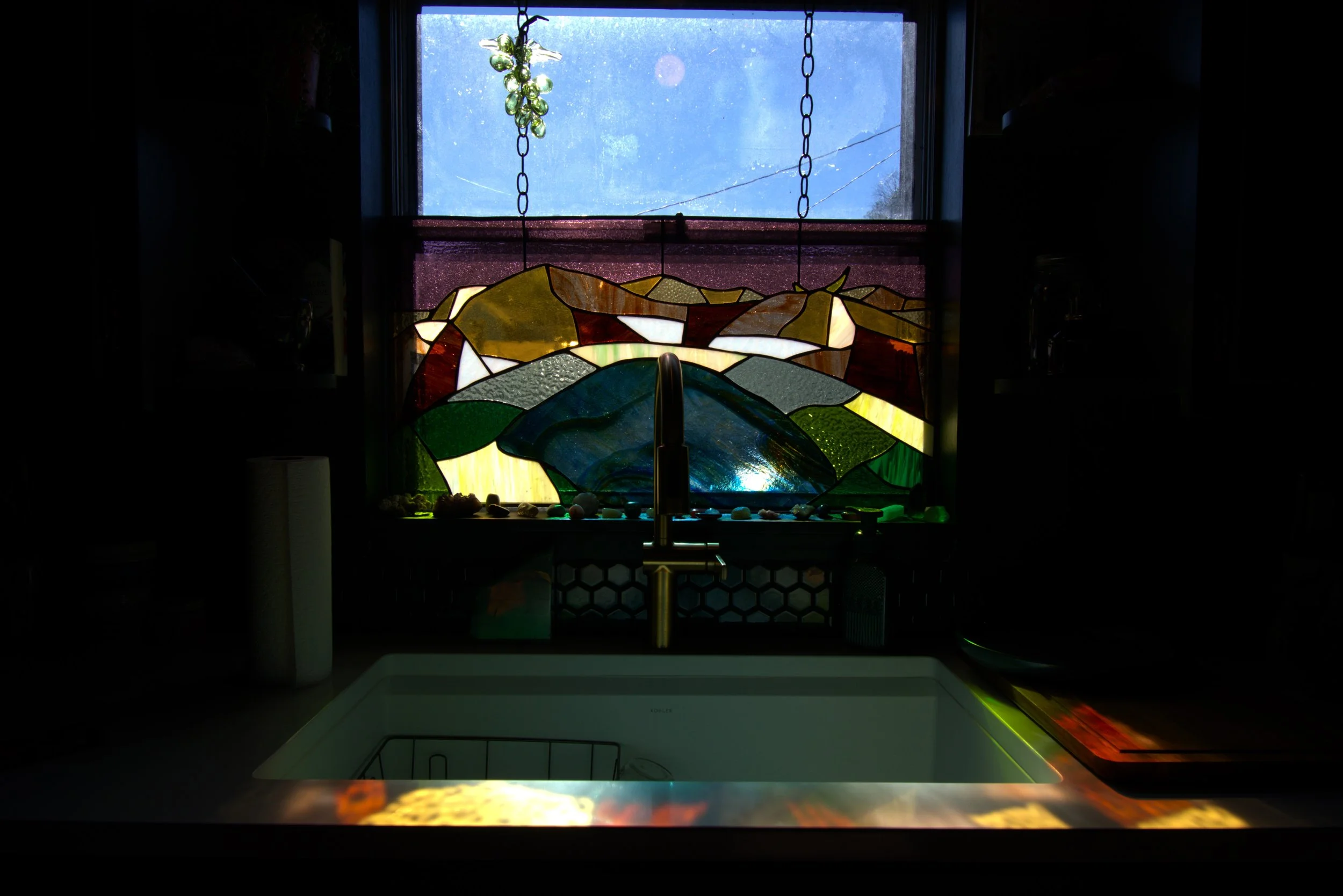A kitchen window with a stained glass design featuring colorful elements, with a small green hanging plant above and a visible sink below the window.