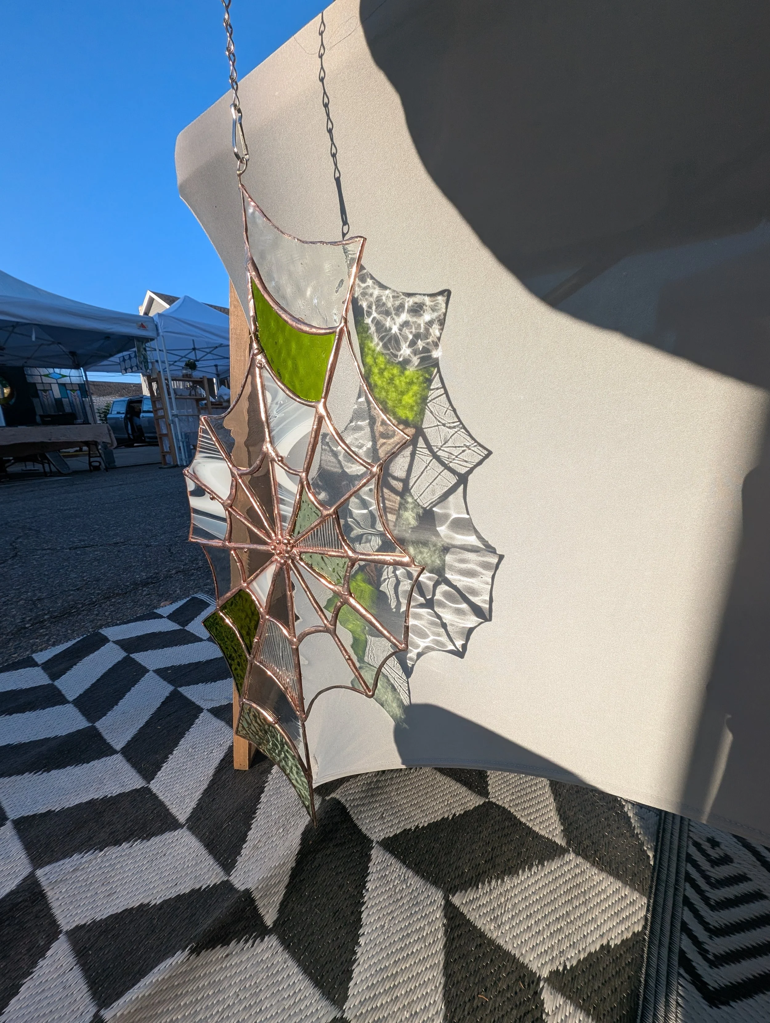 Decorative glass mosaic in the shape of a spider web with green and clear glass segments, attached to a white board at an outdoor craft fair with tents and a parked vehicle in the background.