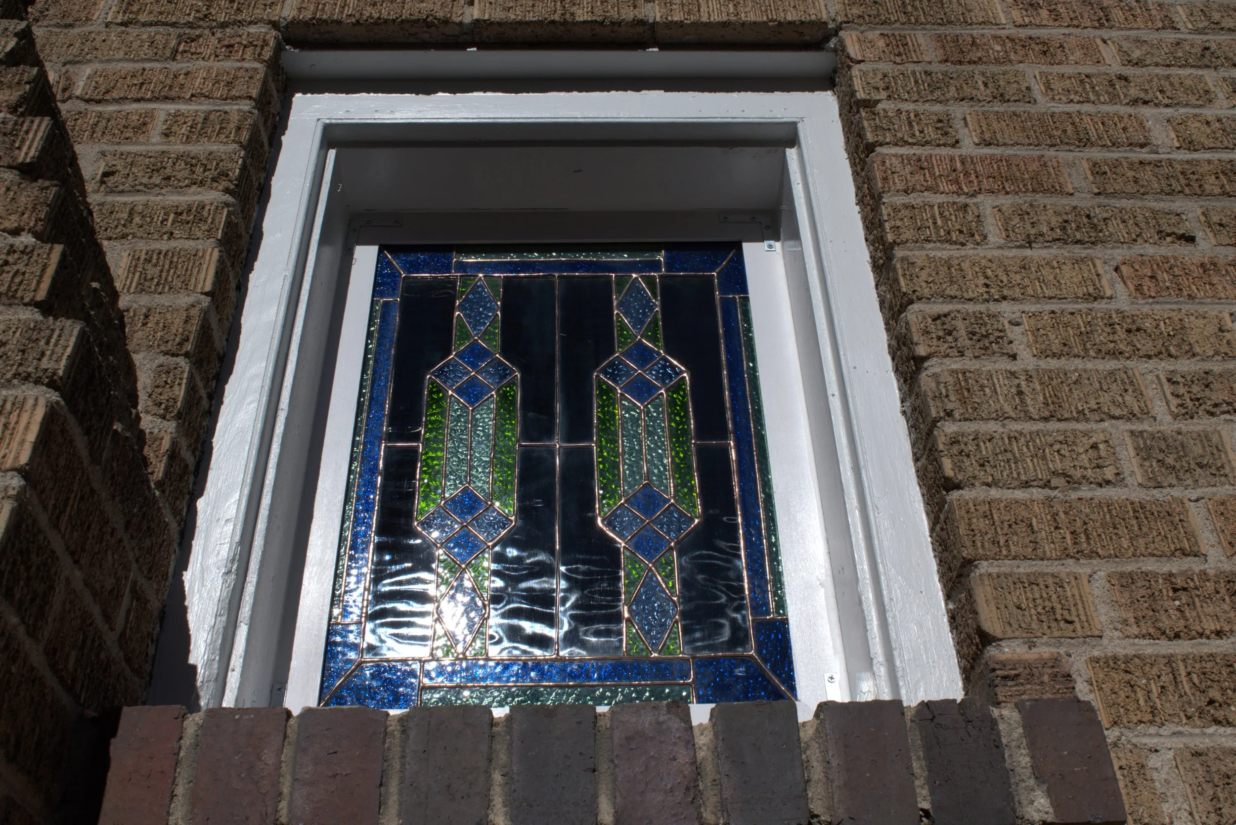 Oriental stained glass window embedded in a brick wall, featuring intricate geometric patterns in blue, green, and clear glass.