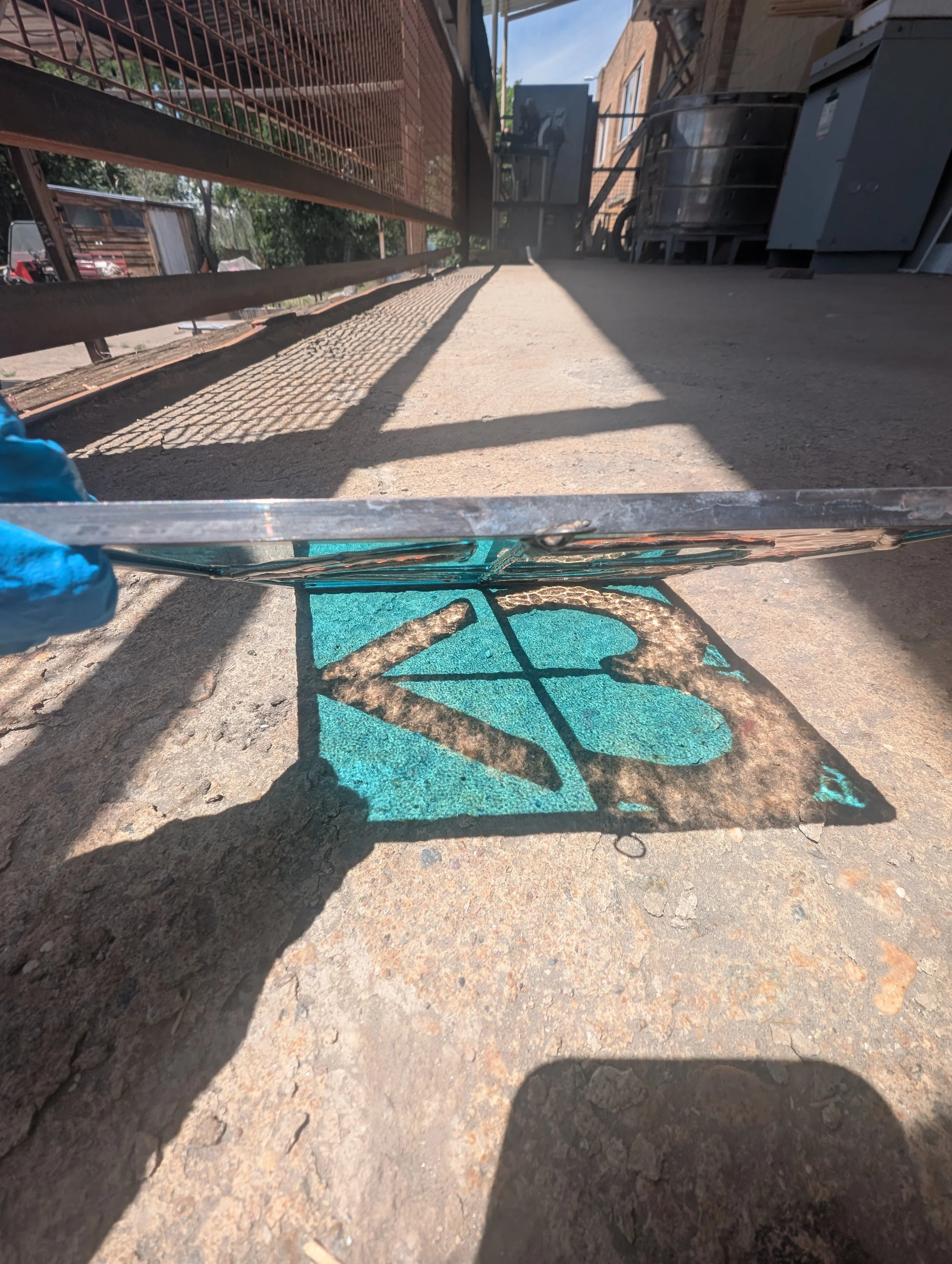 A person wearing a blue glove holding a glass panel over a vintage metal sign with the letters 'NS' on it, casting a shadow on a concrete sidewalk.