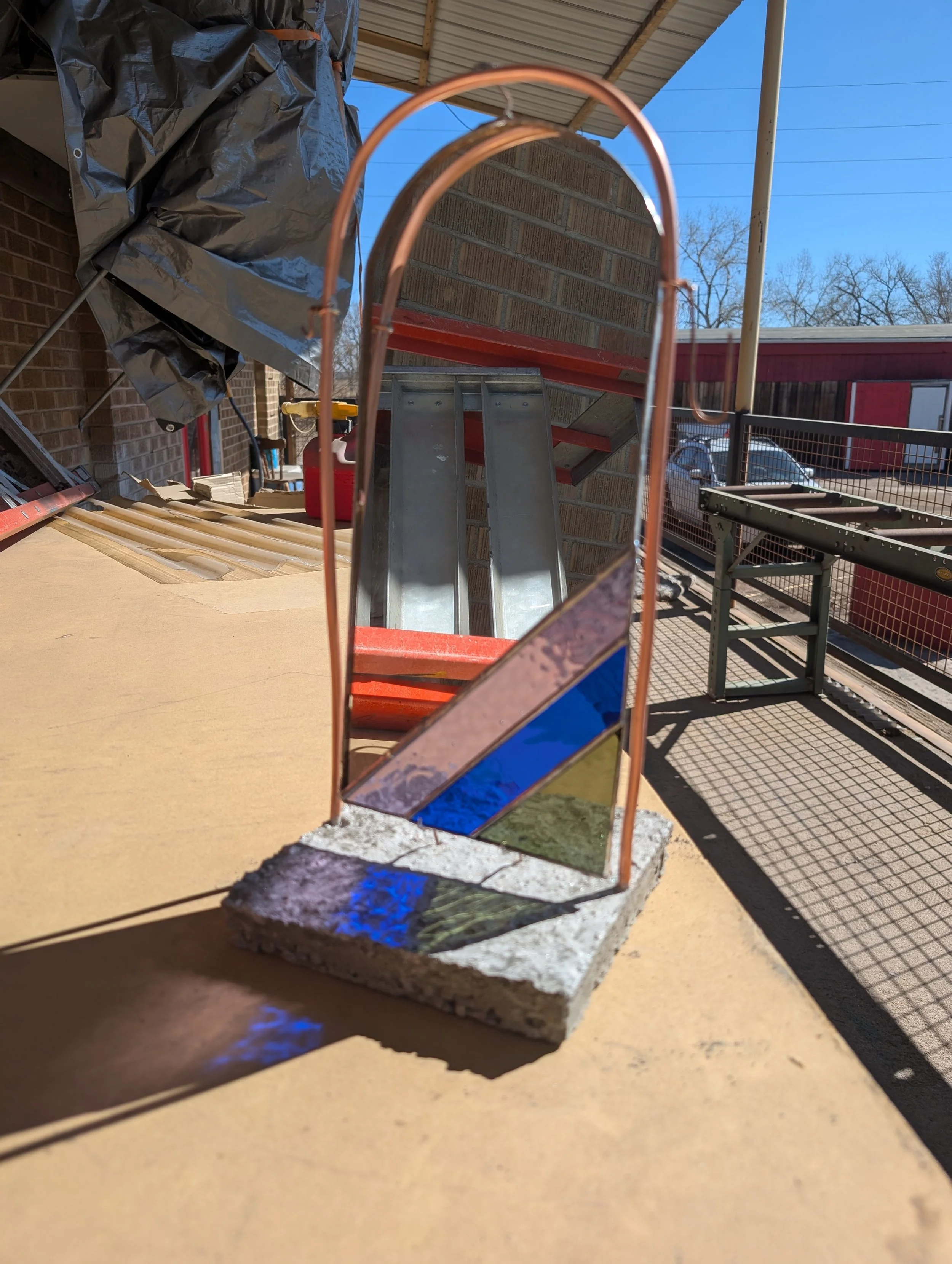 A stained glass art piece with colored glass pieces on a concrete base, casting colorful reflections on a yellow surface outside.